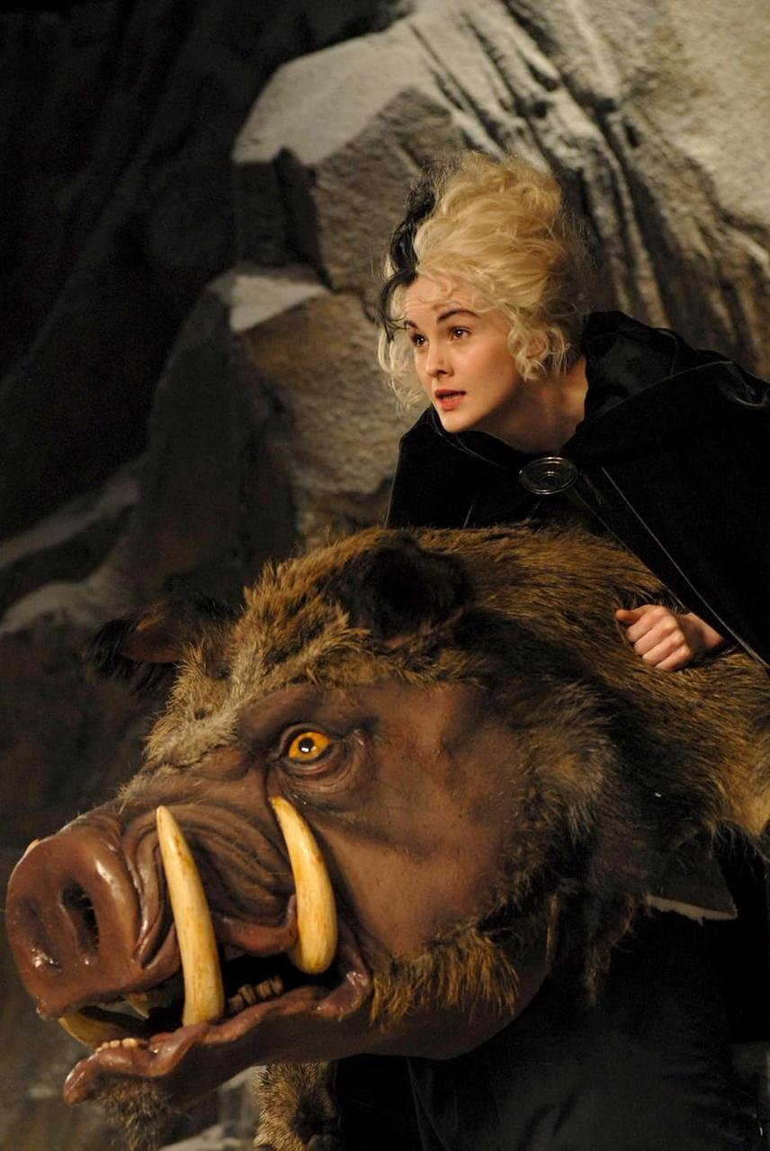 Eine Frau mit schwarz-blondem Haar blickt über einen Wildschweinkopf-Requisit. Sie trägt einen schwarzen Umhang. Der Hintergrund ist eine felsige Höhle.
