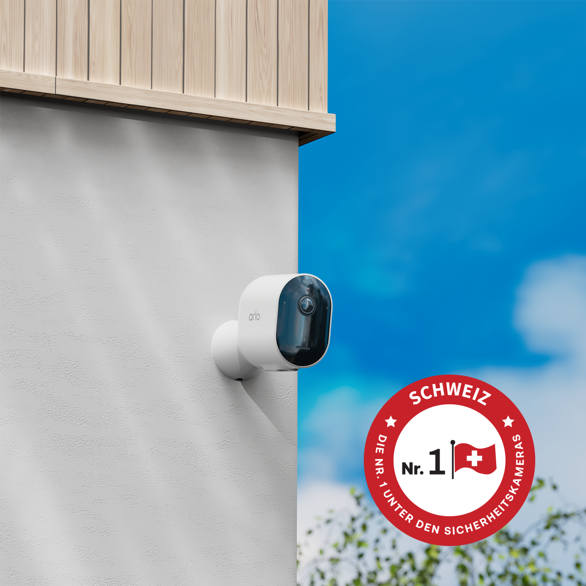 Une caméra de sécurité Arlo blanche est montée sur un mur blanc. Un badge rouge avec le drapeau suisse et du texte est visible.