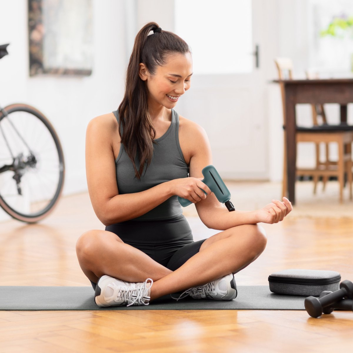 Woman using a massage gun on her arm while sitting on a yoga mat. A bicycle, dumbbells, and a case are in the background.