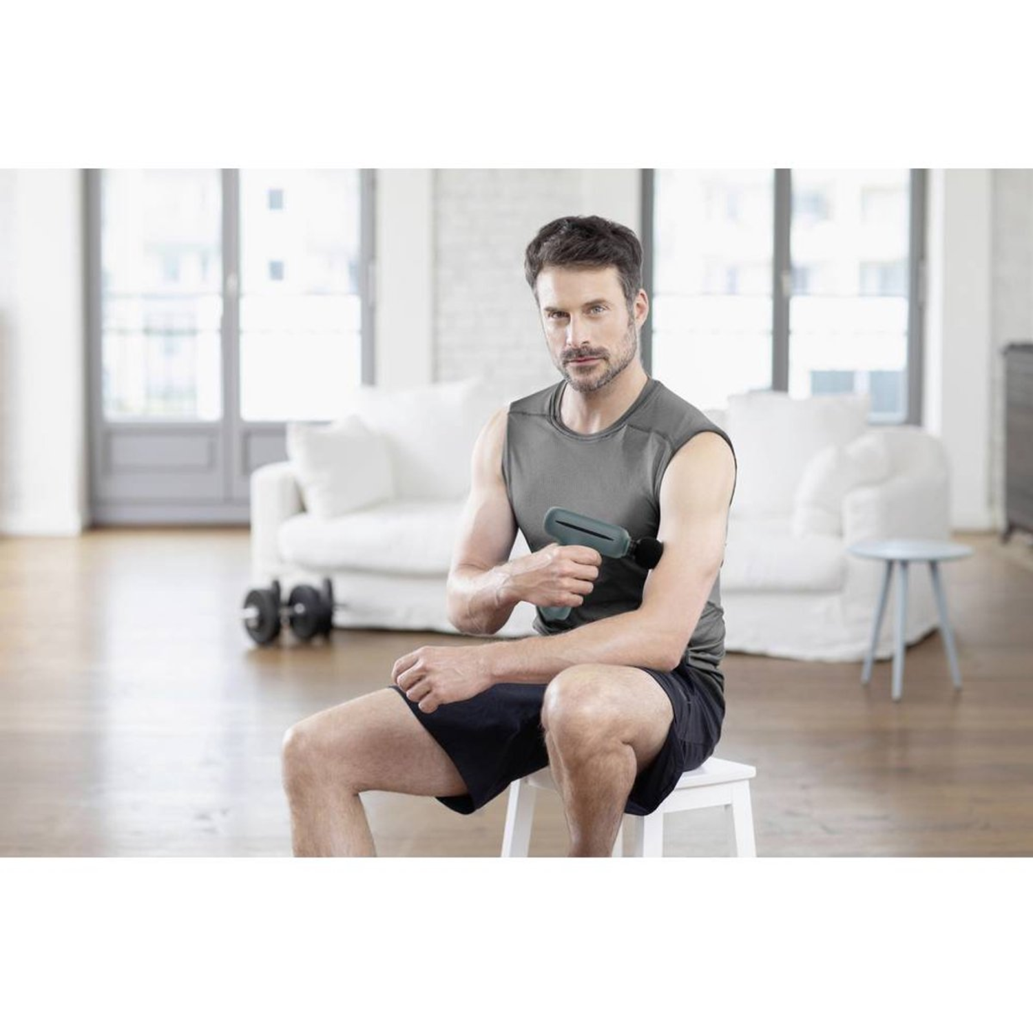 A man sits, using a massage gun on his arm. He's in a room with a white couch, dumbbells, and a stool. He wears shorts and a tank top.