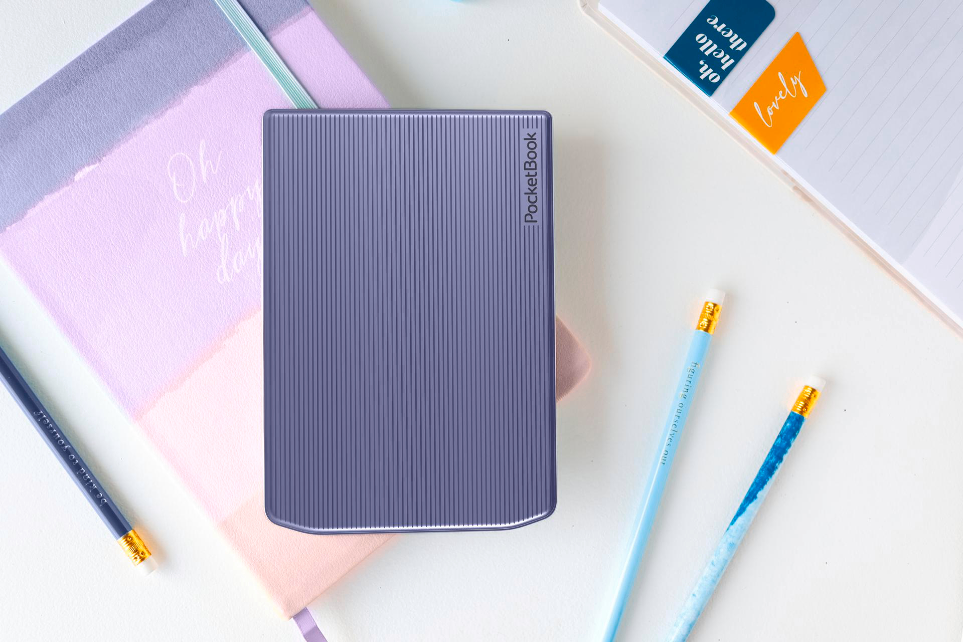 Ein lila E-Reader steht im Mittelpunkt einer weißen Oberfläche, umgeben von Schreibwaren wie Bleistiften und Notizbüchern mit inspirierenden Zitaten.