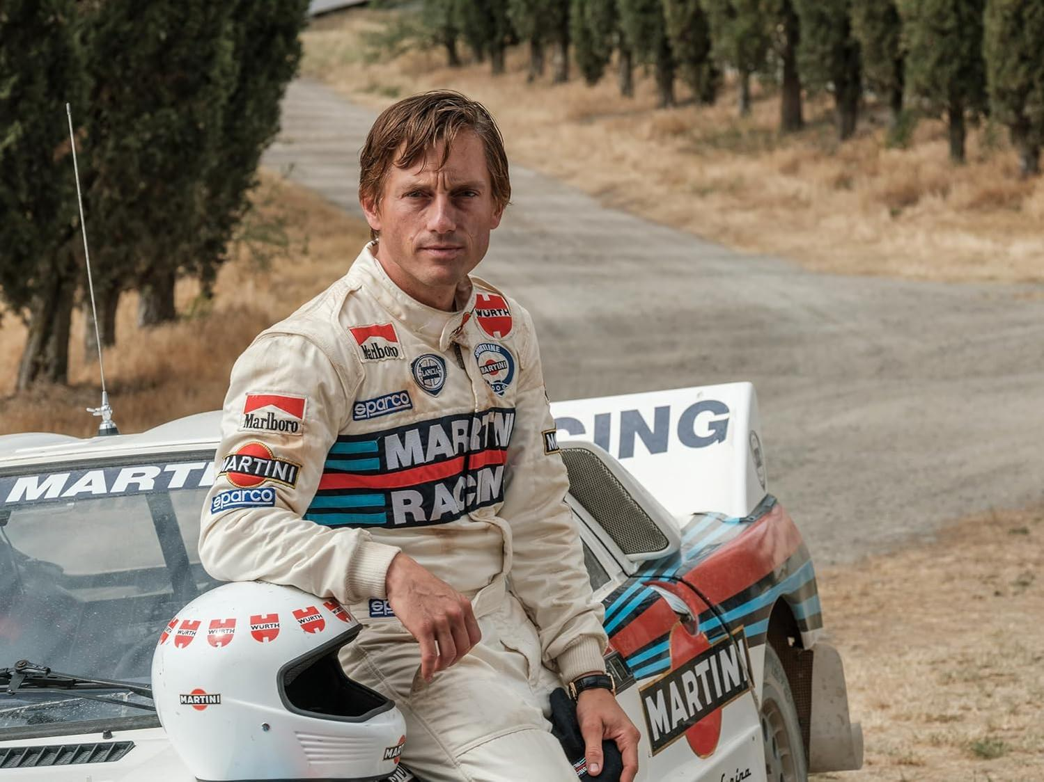 Ein Mann in einem Rennanzug sitzt auf einem Rallyeauto. Das Auto hat Martini Racing-Aufkleber. Ein Helm ist im Vordergrund.