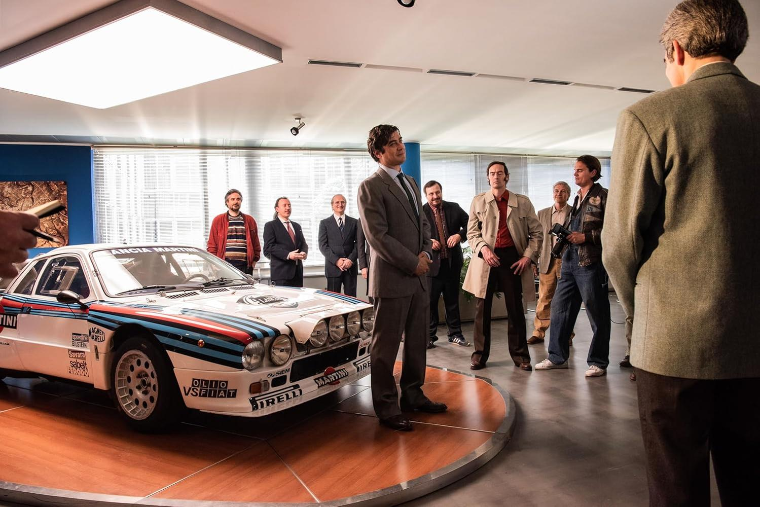 Männer versammeln sich um ein weißes Rallyeauto. Ein Mann im Anzug steht davor. Andere beobachten, einige halten Kameras.