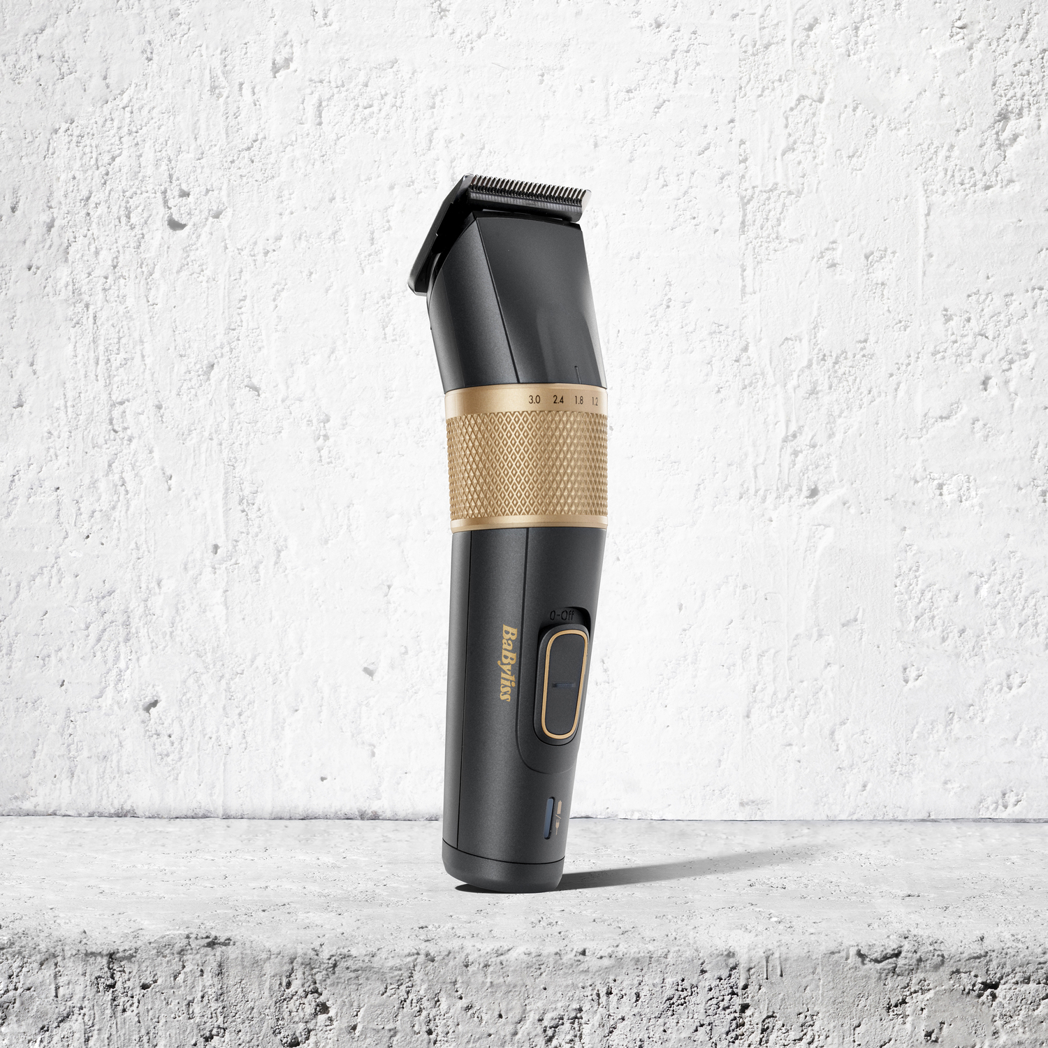 A black and gold Babyliss hair clipper stands on a concrete surface against a textured white wall, shadow cast.