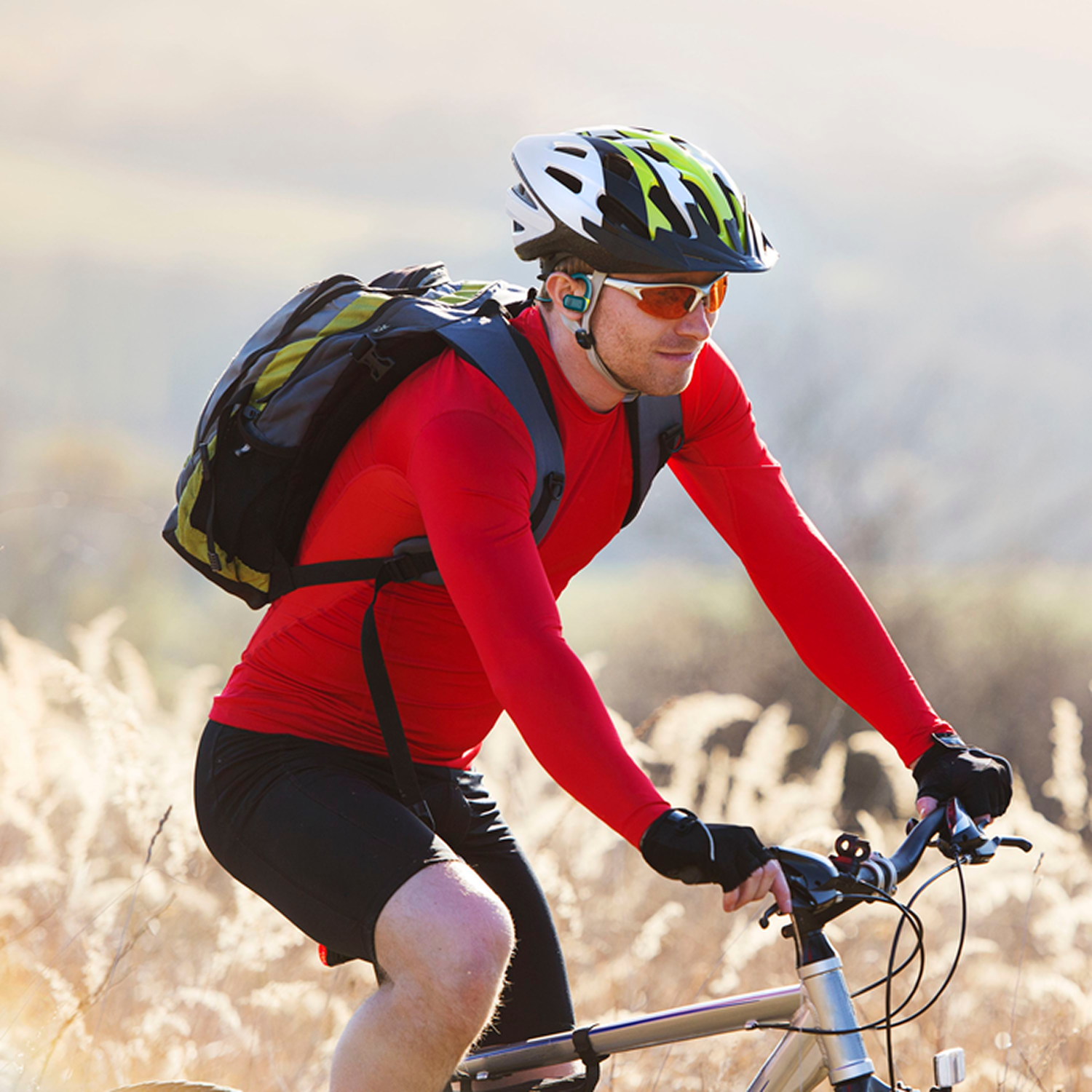 Ciclista con camisa roja y pantalones cortos negros en bicicleta. Lleva casco y gafas de sol. Hierba alta en el fondo.