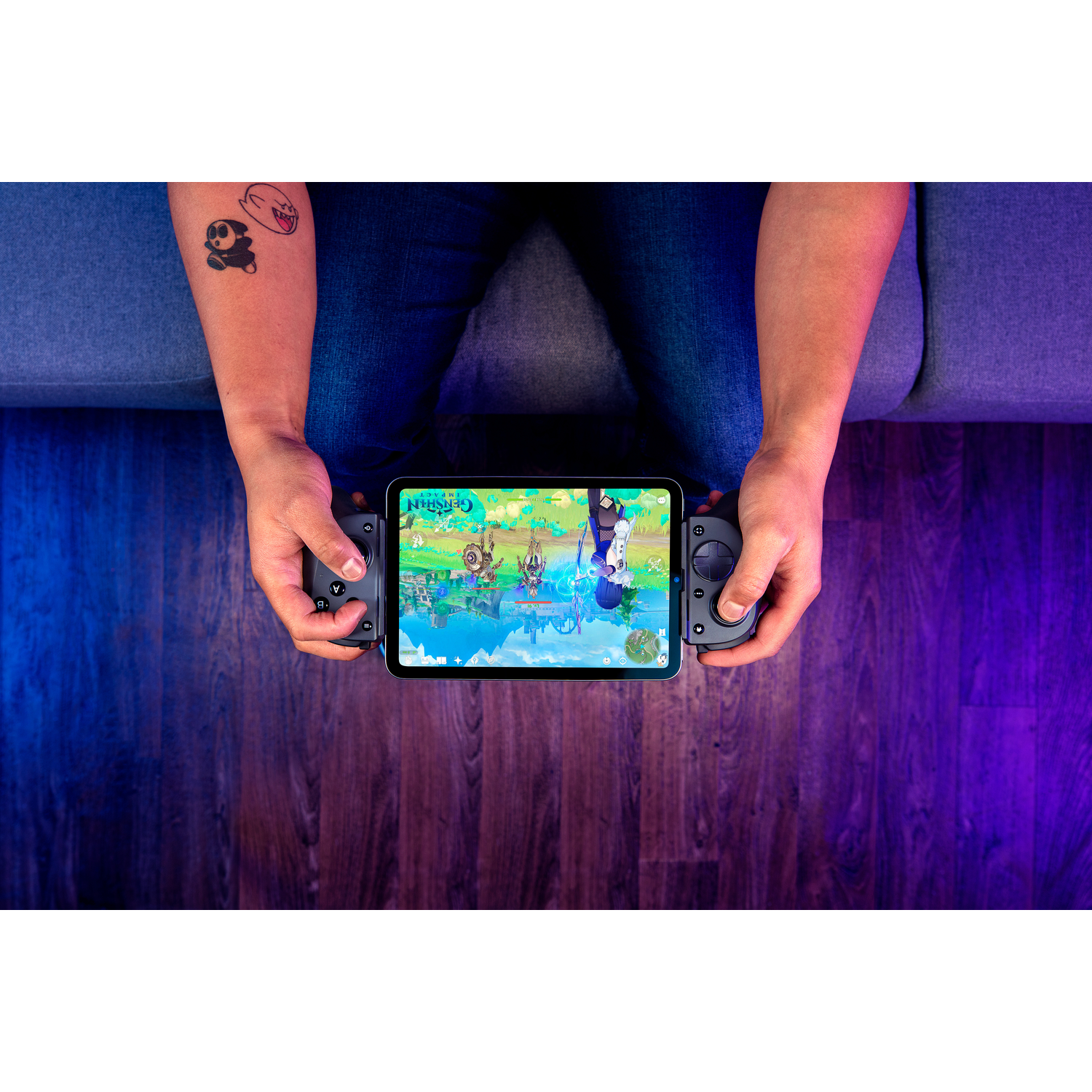 Person holding a tablet with a game displayed, integrated with controllers. Arm has tattoos. Background shows wooden floor and a sofa.