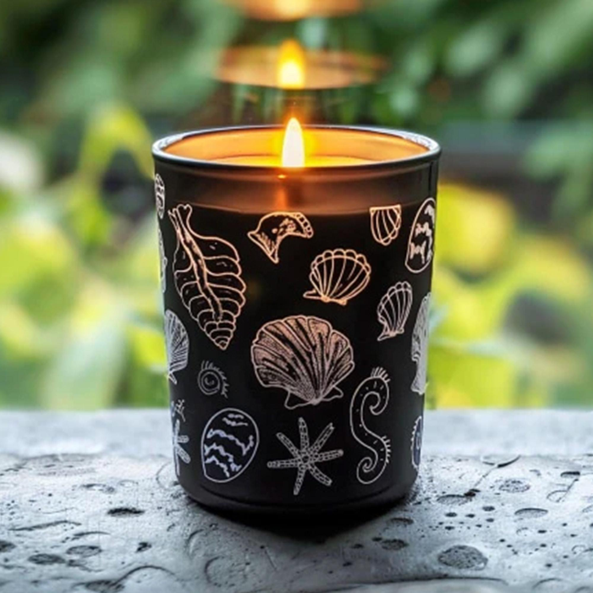 Eine brennende schwarze Kerze mit weißen Muscheldesigns. Sie steht auf einer Betonoberfläche, mit verschwommenem grünem Hintergrund.