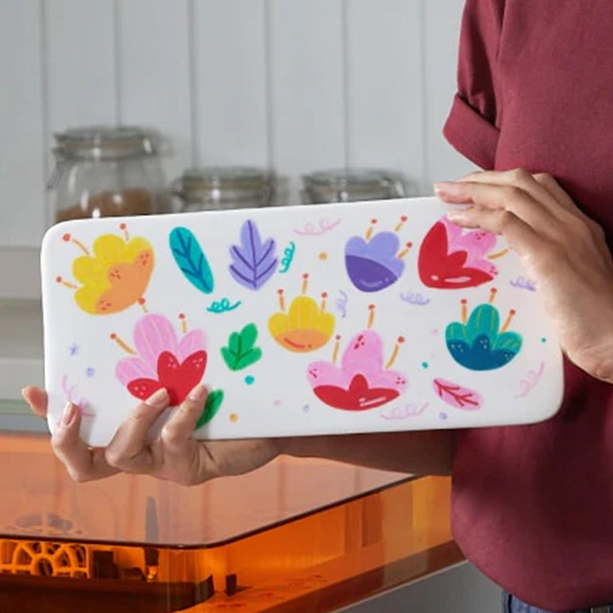 Eine Person hält ein weißes Tablett mit bunten Blumendesigns. Der Hintergrund zeigt Gläser und eine Arbeitsplatte.