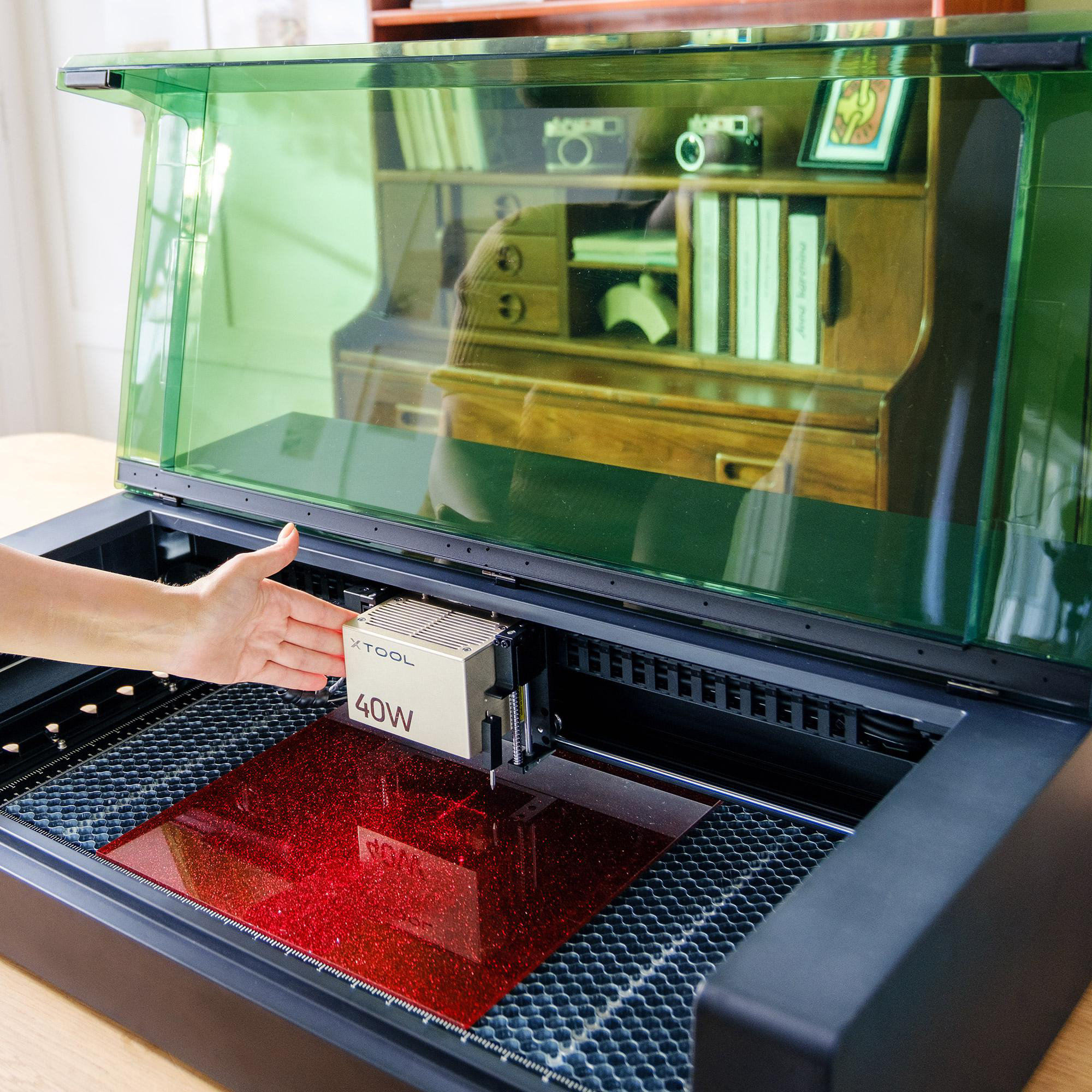 Die Hand einer Person in der Nähe eines Laserschneiders mit grüner Abdeckung. Die Maschine hat eine rote Oberfläche und einen silbernen 40W-Laserkopf.