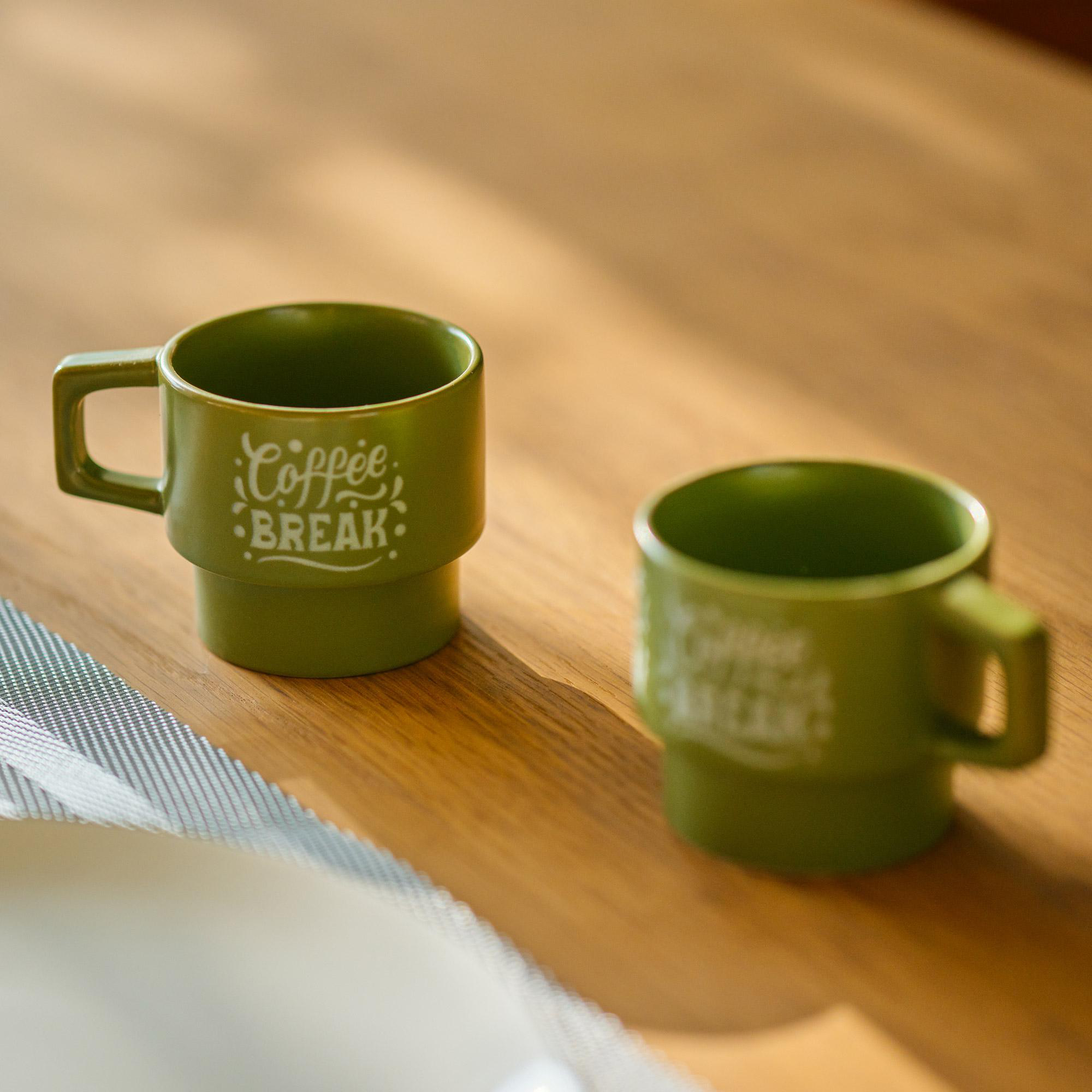 Zwei olivgrüne Tassen auf einem Holztisch. Auf einer Tasse steht "Coffee Break". Weiches Licht beleuchtet die Szene.