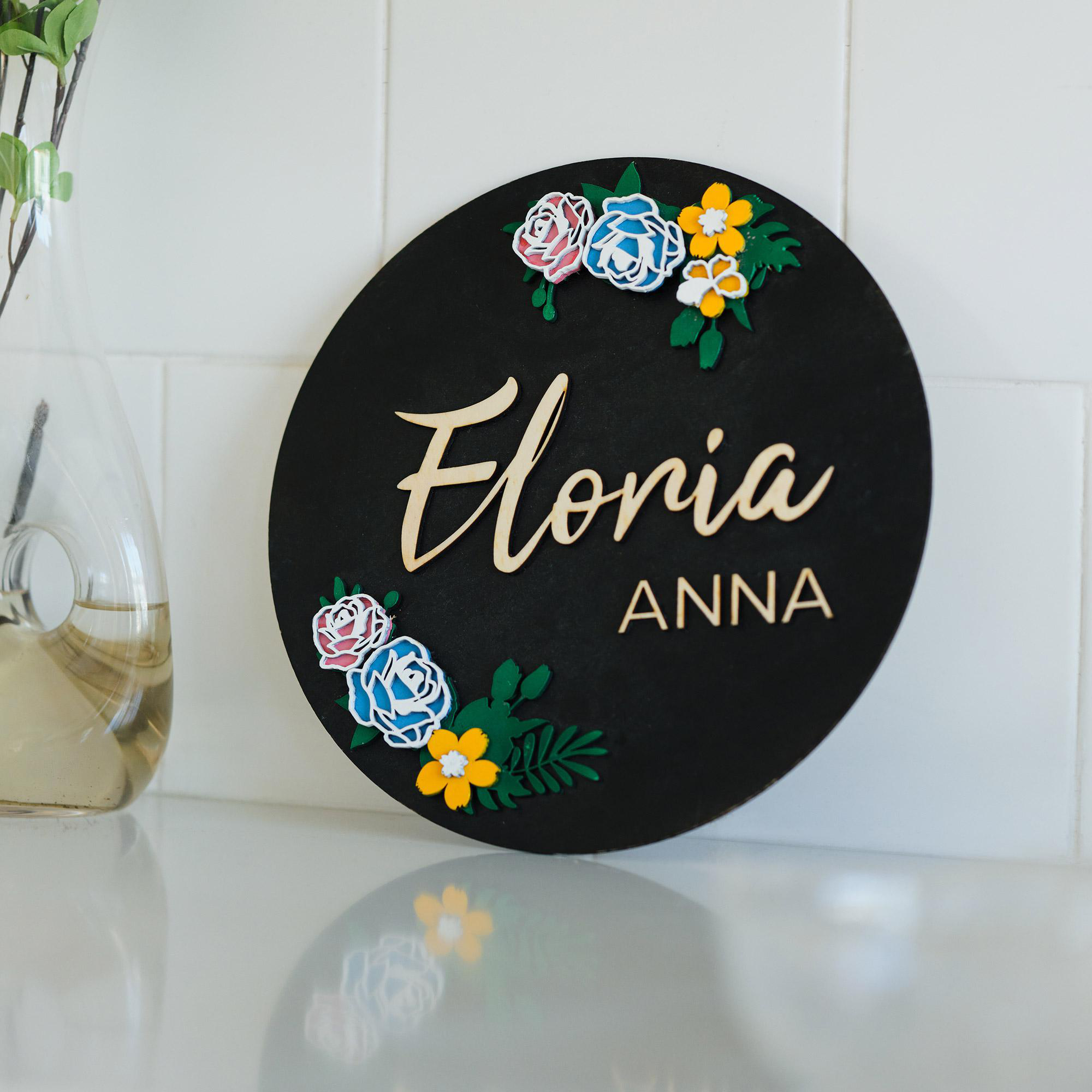 Ein rundes schwarzes Schild mit dem Namen 'Eloria Anna', floralen Akzenten. Links eine Vase mit grünen Blättern.