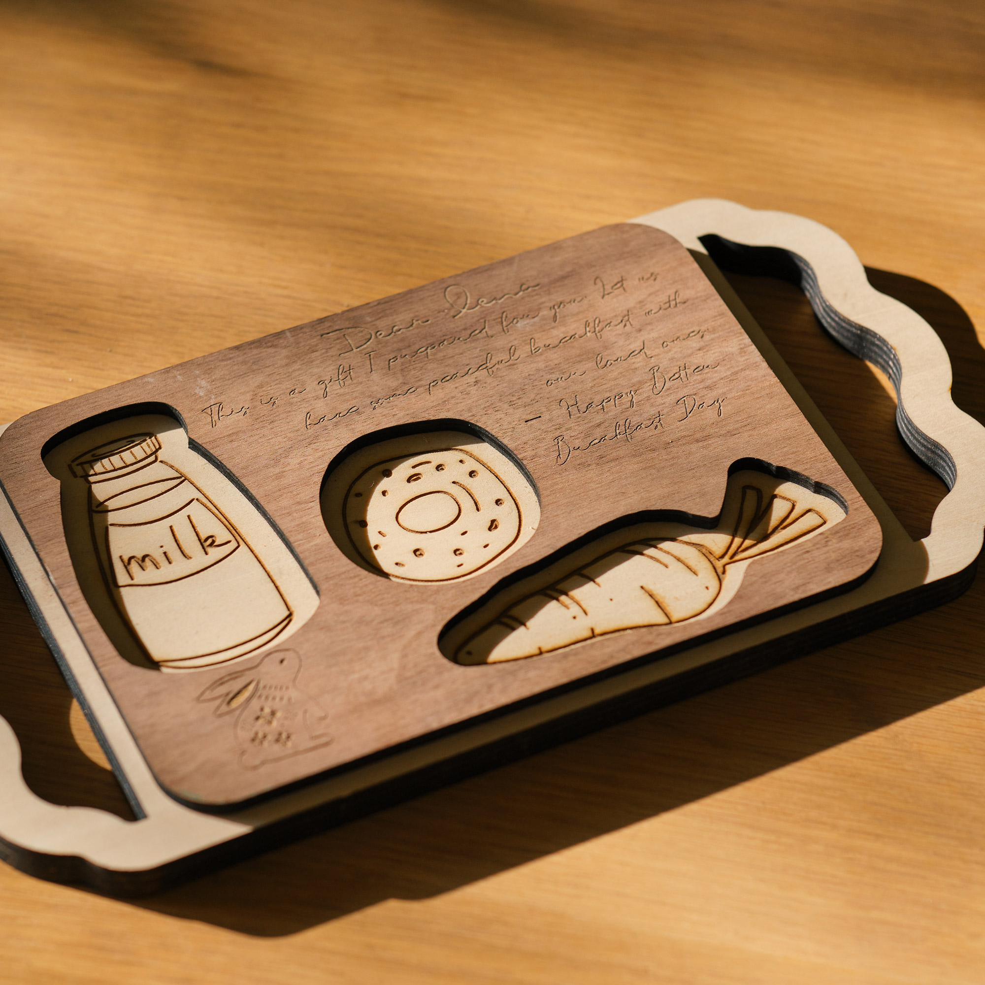 Frühstückstablett aus Holz mit Milch-, Donut- und Karottenausschnitten. Text: Liebe Lena, Happy Breakfast Day. Tablett auf Holztisch.
