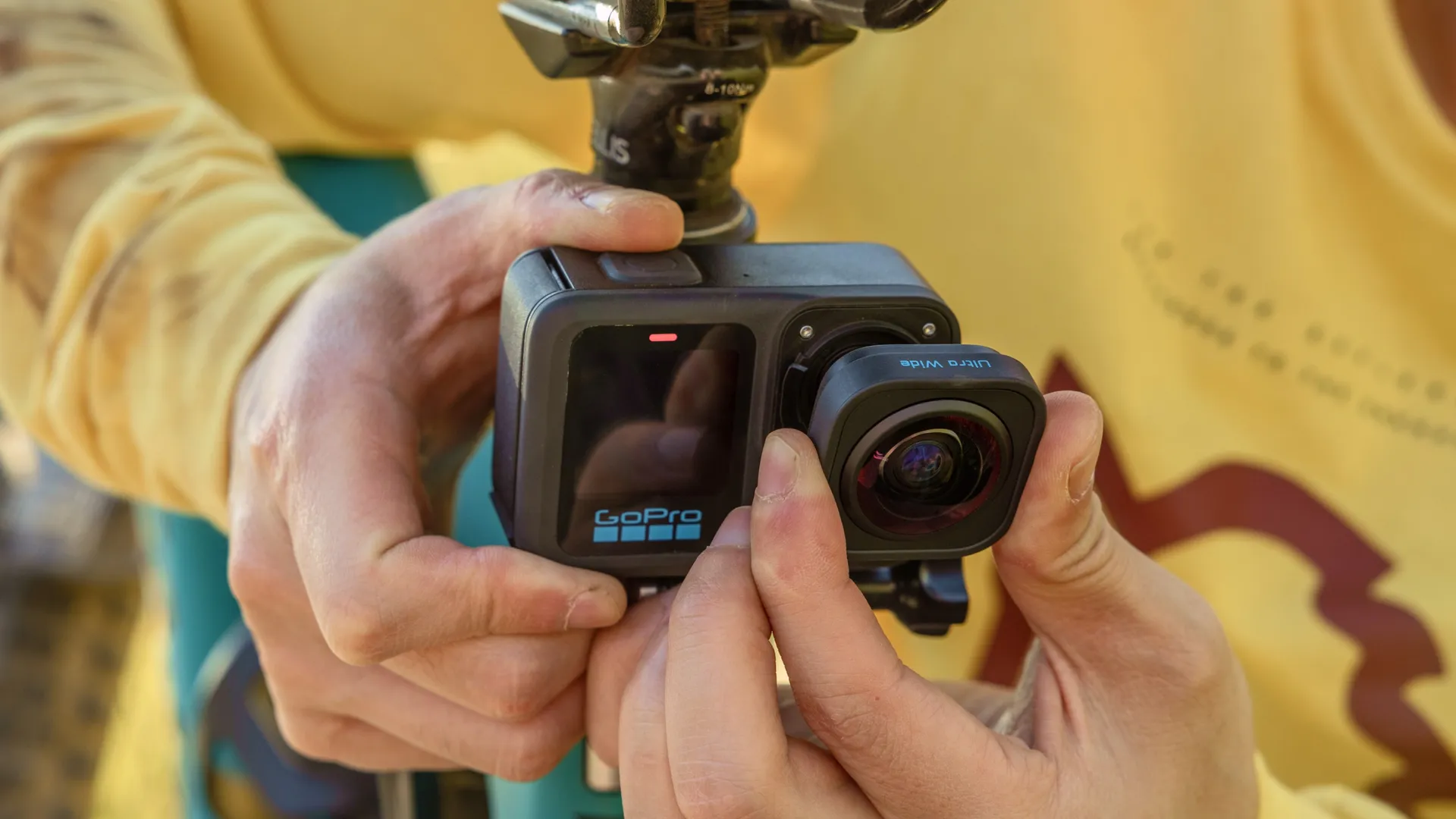Egy személy egy GoPro kamerát tart, beállítva az objektívet. A kamera fekete, nagylátószögű objektívvel. A háttér egy sárga ing.