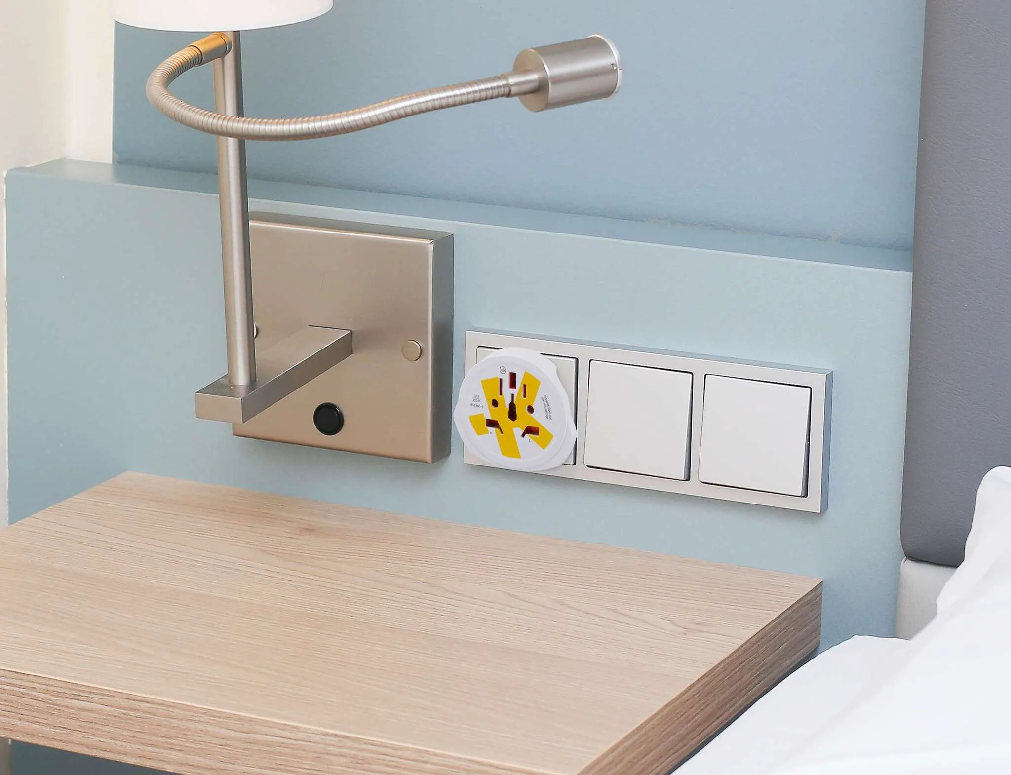 Scena del comodino con lampada, prese e tavolo in legno. Letto bianco a destra. La parete è azzurra.