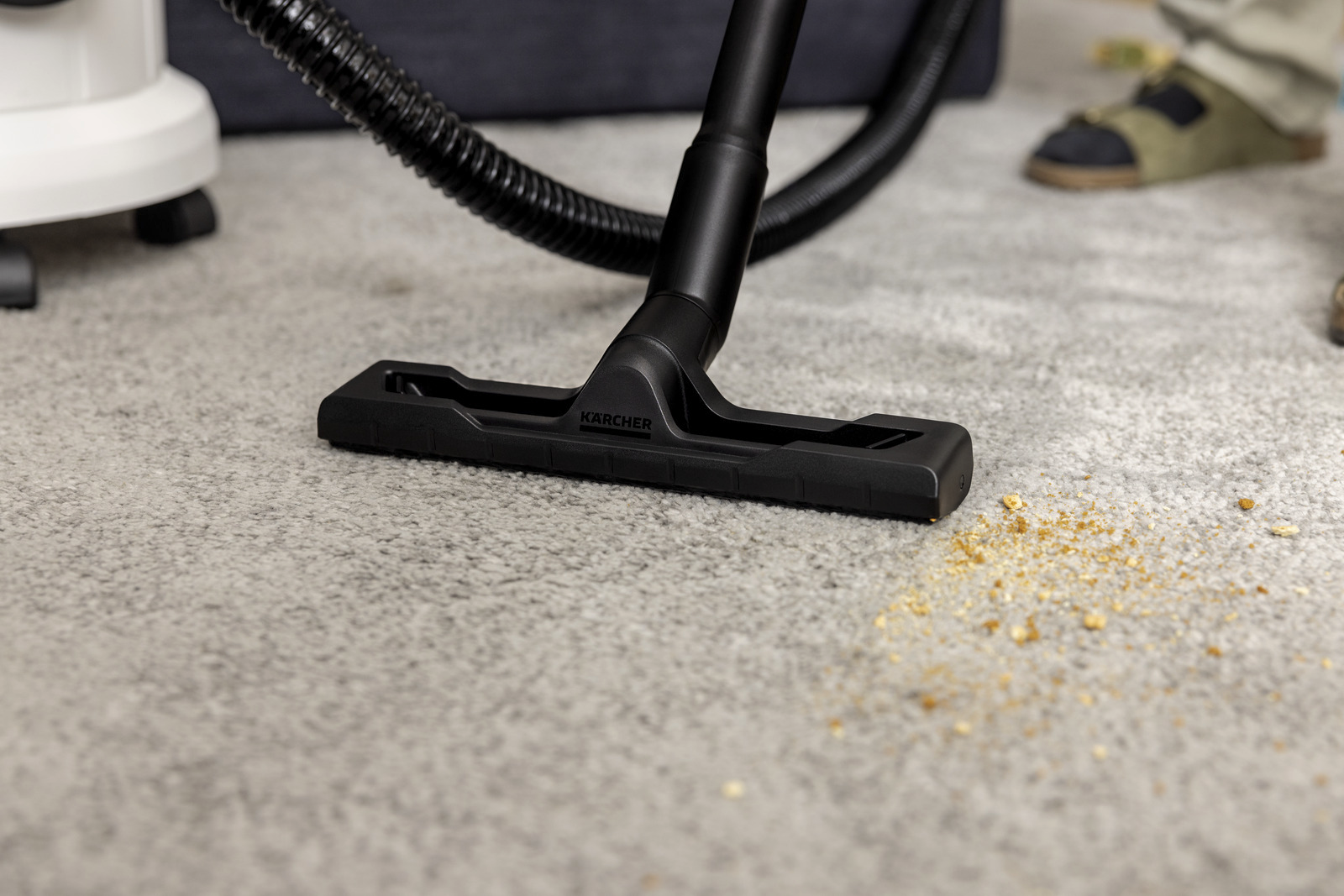 Egy fekete Karcher porszívófej szürke szőnyegen van, morzsákkal és fekete tömlővel. Egy ember lába látható.