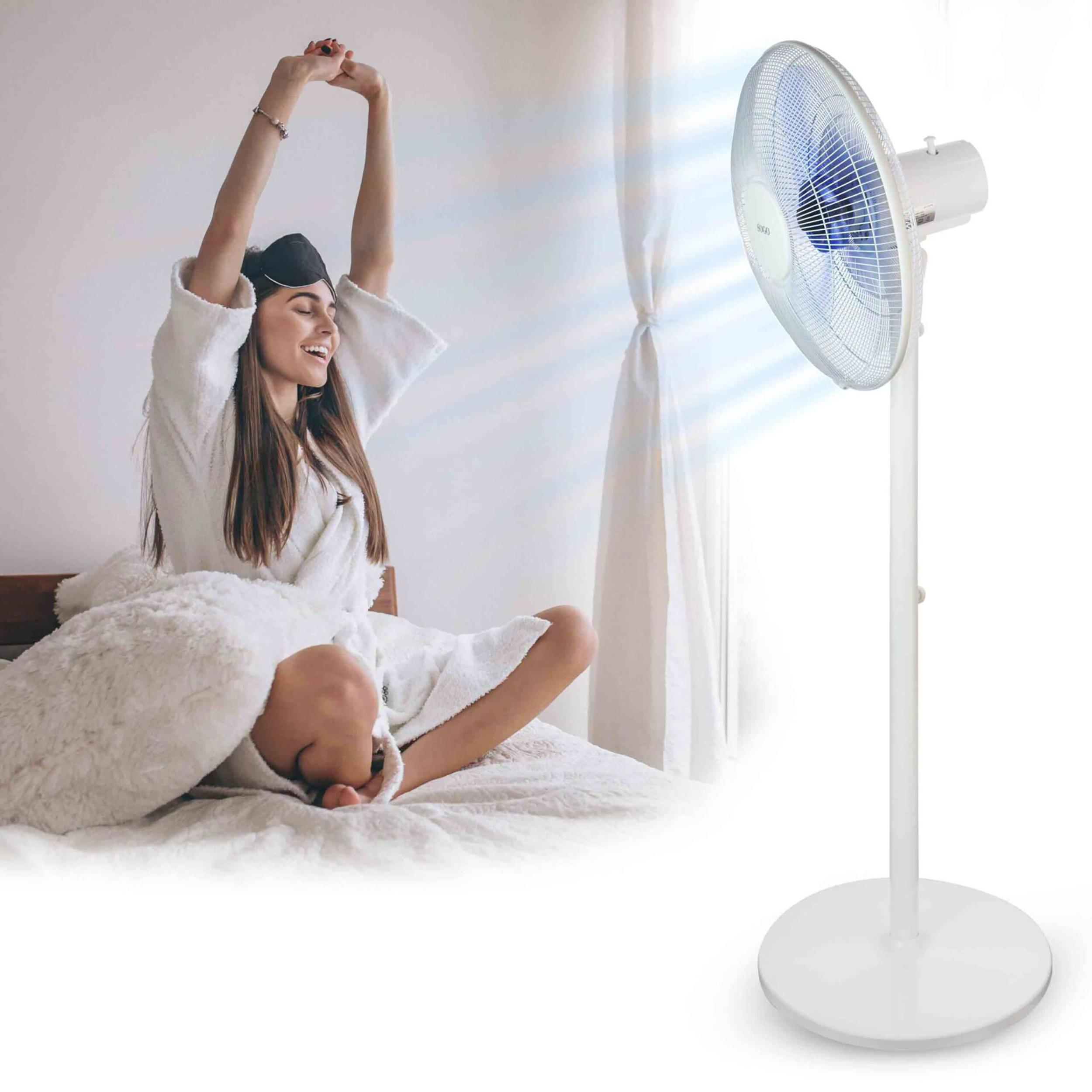 Eine Frau im weißen Bademantel dehnt sich im Bett, mit Schlafmaske. Rechts steht ein weißer Ventilator.