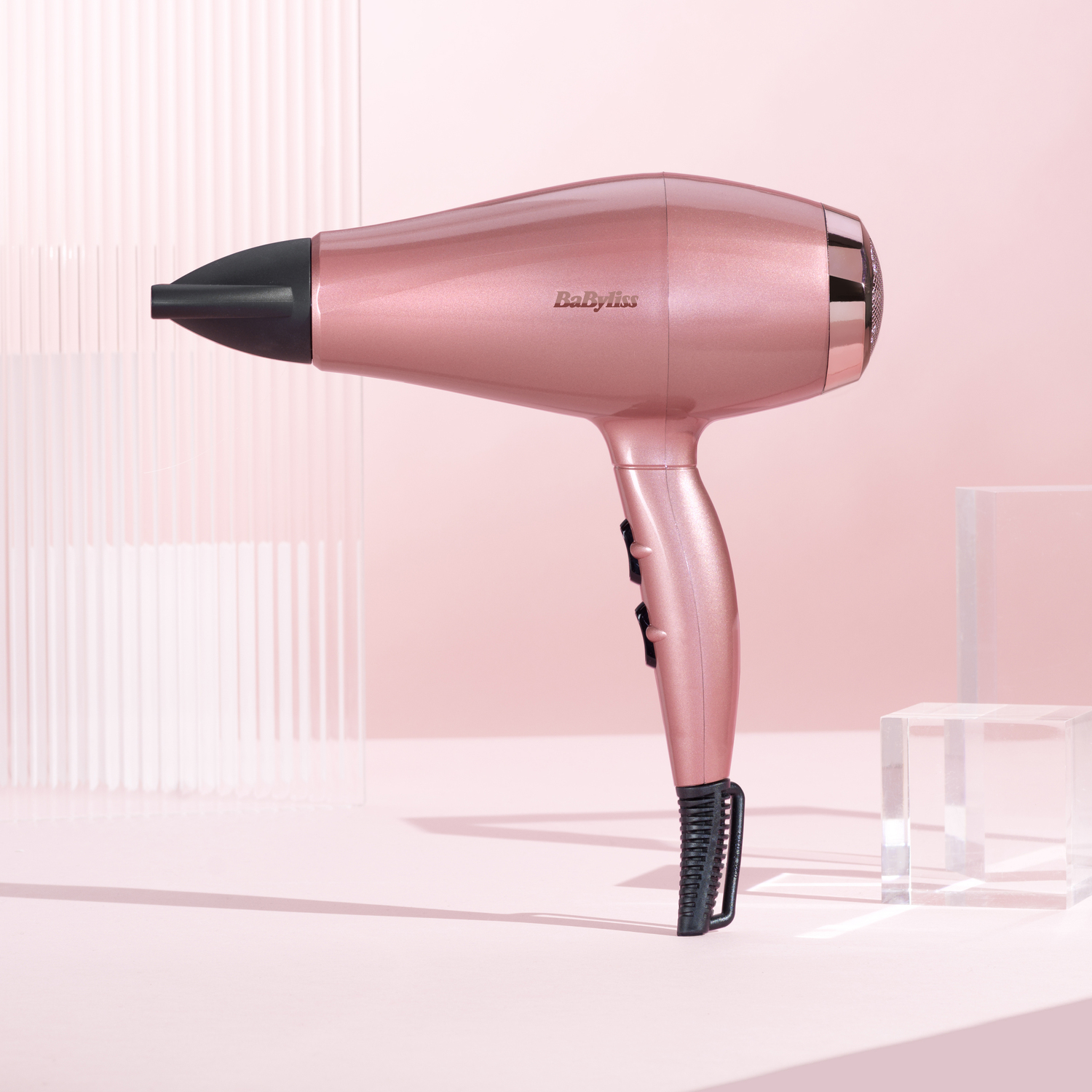 Een roze Babyliss föhn op een roze oppervlak. De achtergrond toont een roze muur en heldere geometrische blokken.