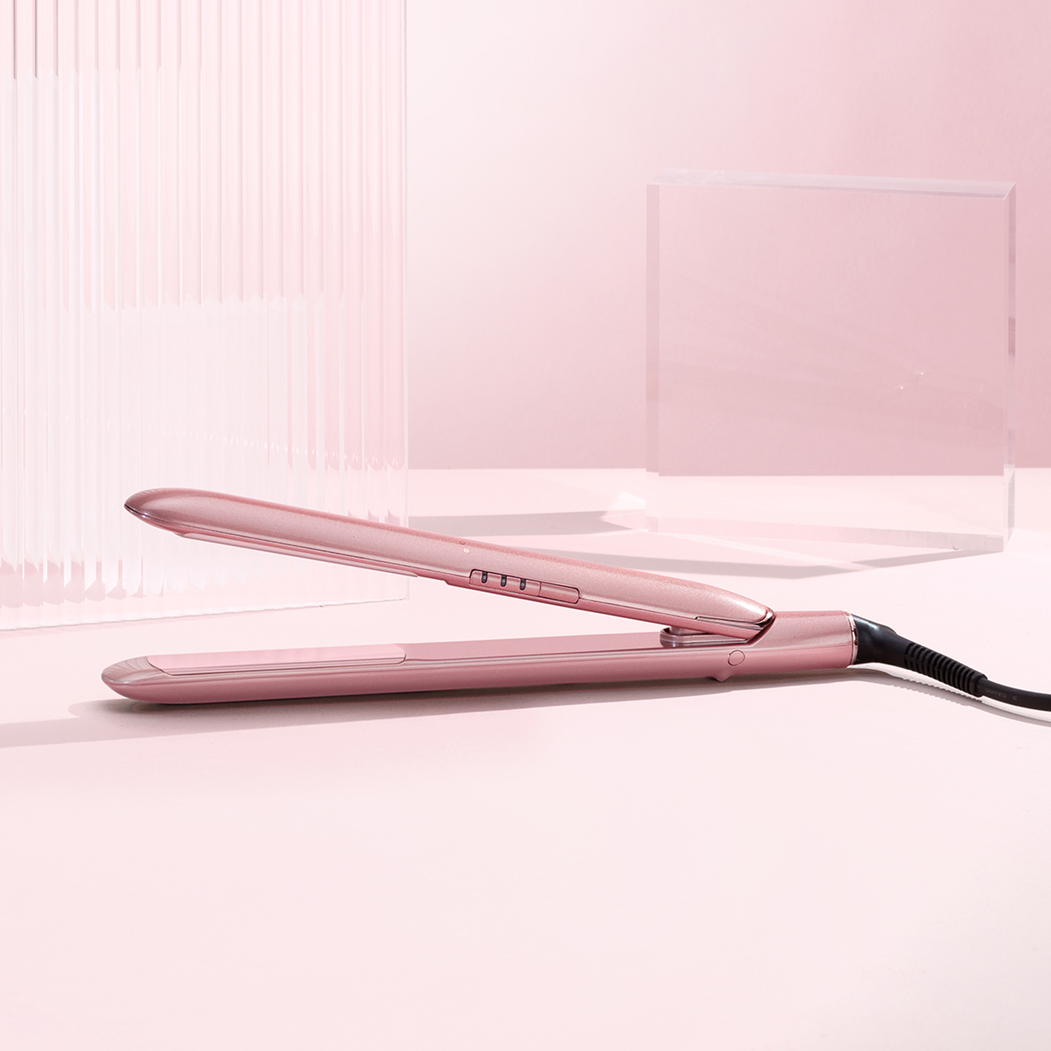 Pink hair straightener on a pink surface. The straightener is open, with a black cord. Clear blocks in the background.