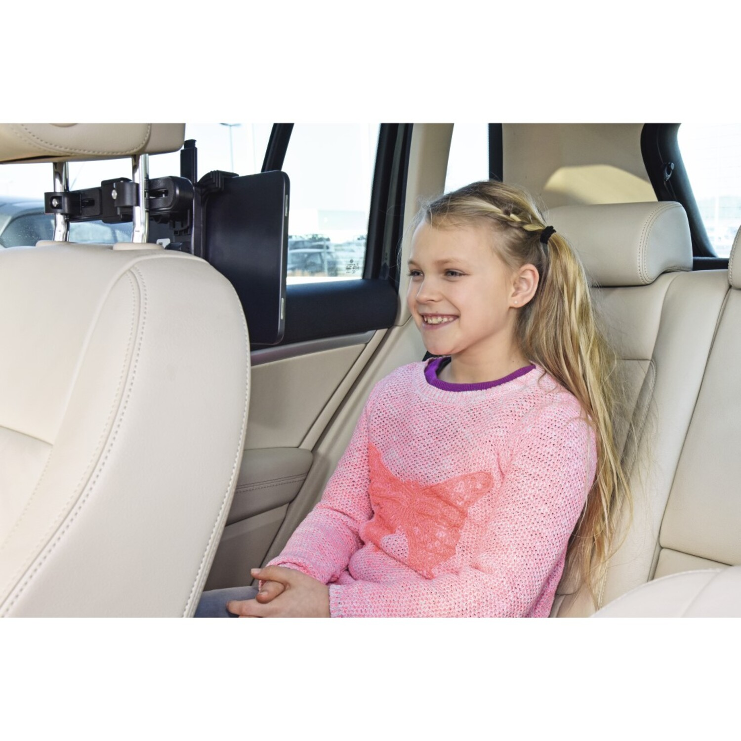 Une fille sourit dans une voiture. Une tablette est fixée à l'arrière du siège avant. La fille porte un pull rose.