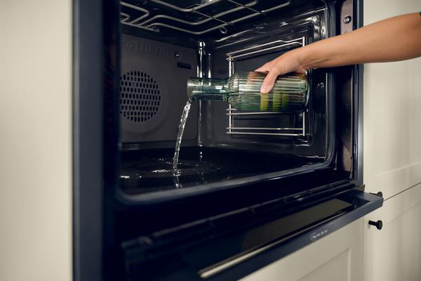 Eine Hand gießt Wasser aus einer grünen Glasflasche in einen Ofen. Wasser fließt in das dunkle Ofeninnere.