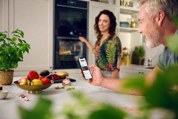 Ein Mann hält ein Telefon in einer Küche. Eine Frau steht in der Nähe eines Ofens. Obst und Gemüse liegen auf der Theke.