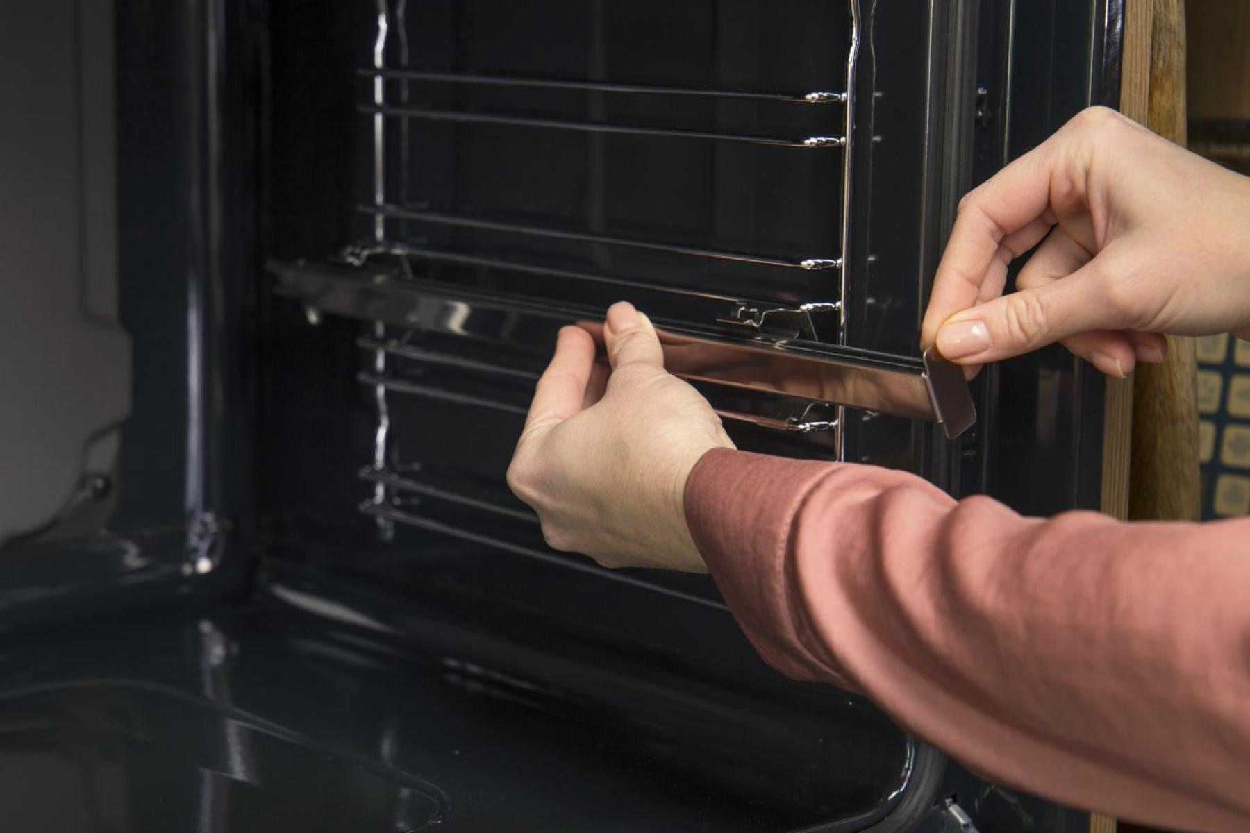 Osoba instalująca ruszt piekarnika. Dłonie trzymają ruszt i mały kawałek metalu wewnątrz piekarnika. Widoczna czerwona koszula.