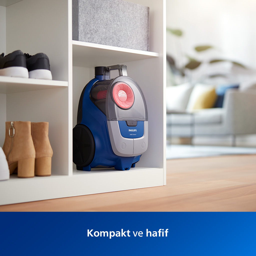 Mavi bir Philips elektrikli süpürgesi beyaz bir dolapta duruyor. Ayakkabılar ve gri bir kutu raflarda. Arka plan bulanık.