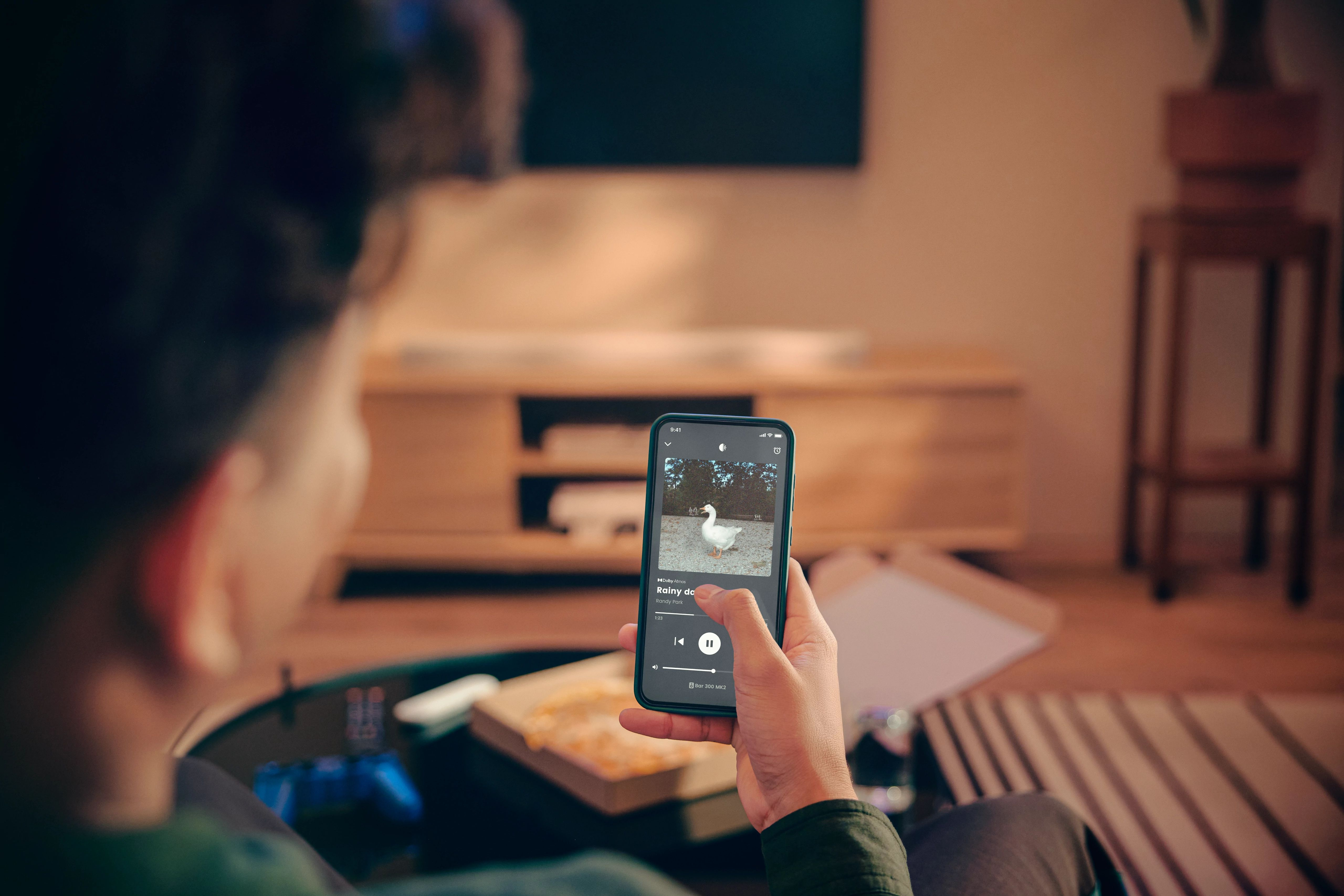 Müzik çalan telefon tutan kişi. Ekran, bir kaz gösteriyor. Arka plan TV, masa ve mobilyaları içerir.