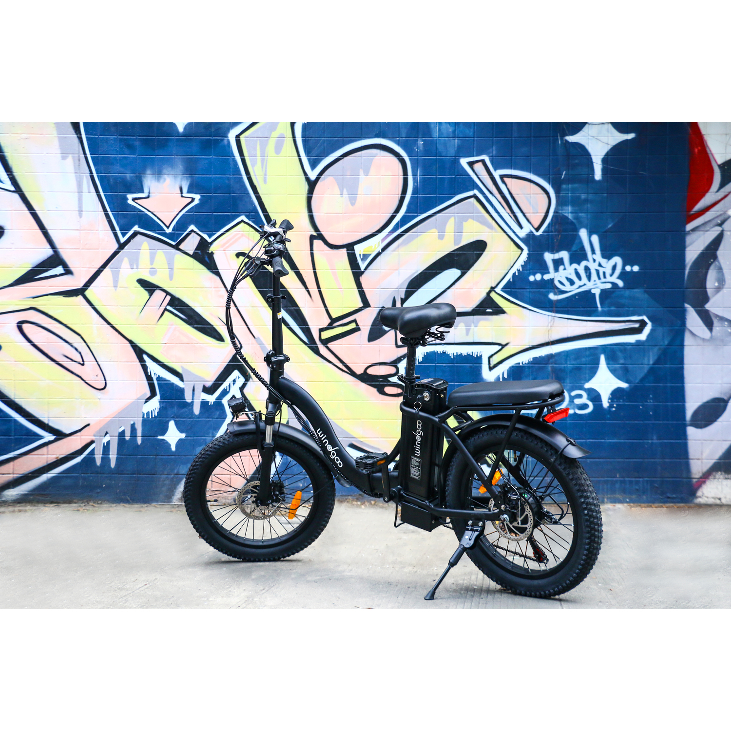 Vélo électrique noir garé sur du béton. Graffiti en arrière-plan. Le vélo a des pneus épais et une batterie.