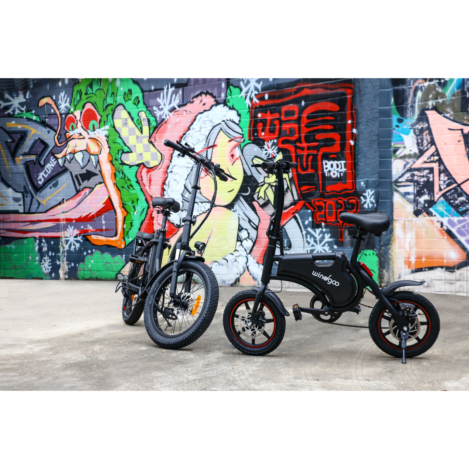 Deux vélos électriques noirs garés sur du béton, contre un mur de briques recouvert de graffitis. Un vélo a des accents orange.