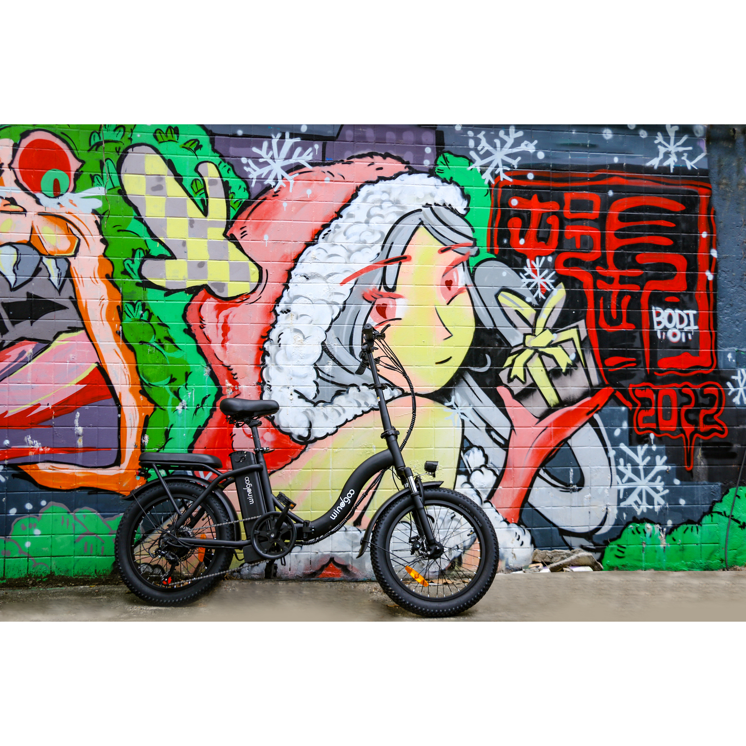 Vélo électrique noir garé devant un mur couvert de graffitis représentant le Père Noël et des flocons de neige. Le vélo a de gros pneus.