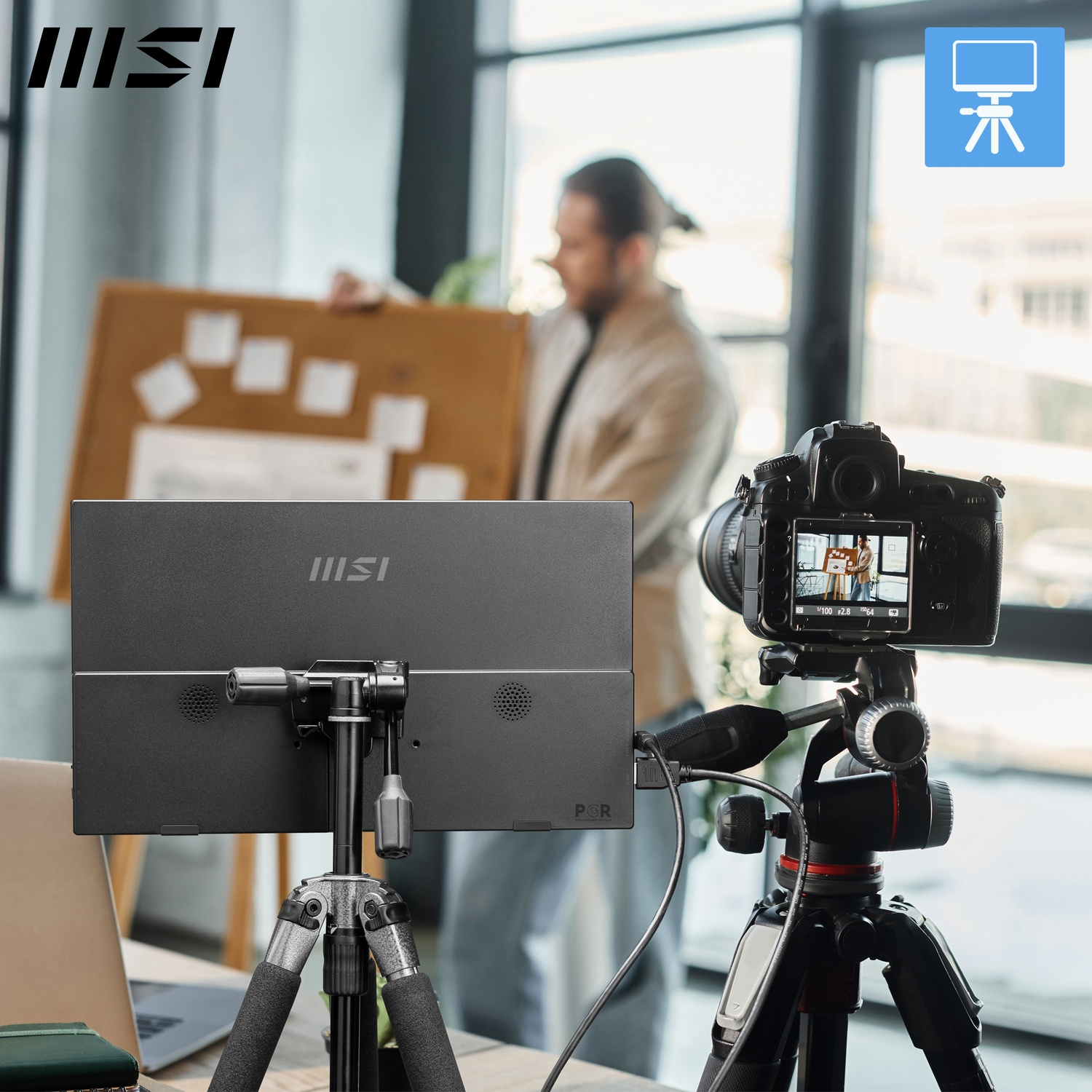 Une configuration vidéo avec un moniteur sur trépied, une caméra et une personne en arrière-plan tenant un tableau de présentation.