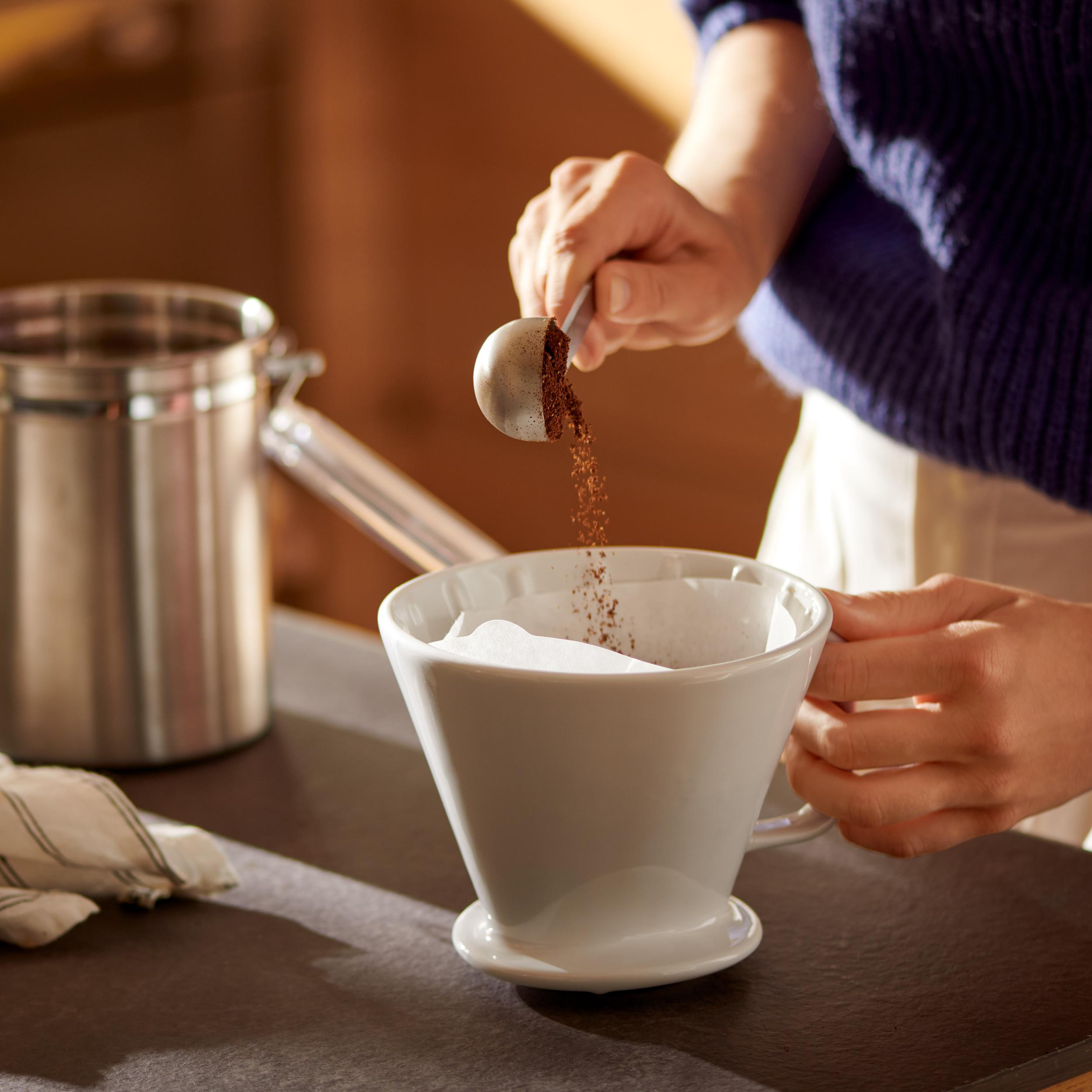 Eine Person gießt Kaffeepulver aus einem Schöpfer in einen weißen Filter. Ein Edelstahlkessel ist im Hintergrund.
