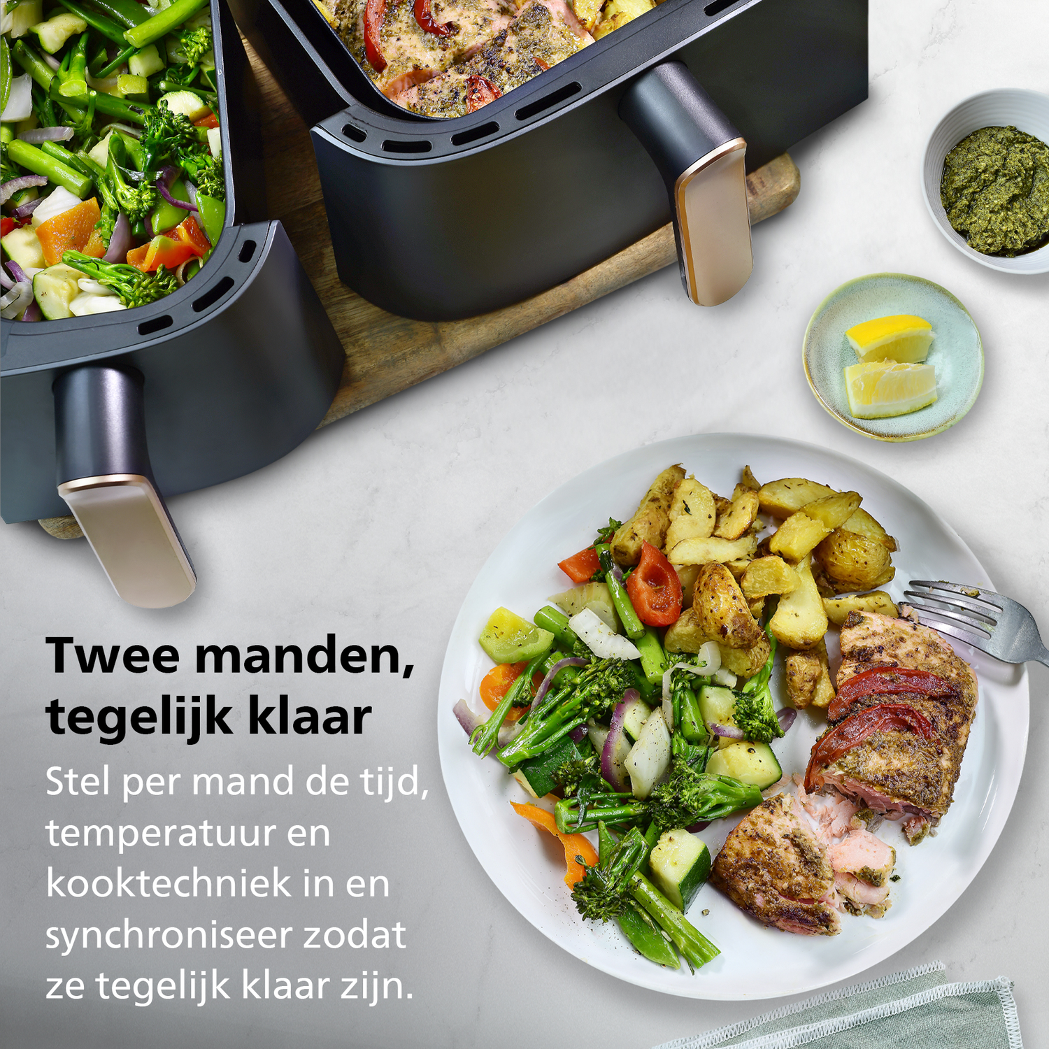 An air fryer with two baskets, food on a plate with vegetables and potatoes, pesto, and lemon slices are visible.