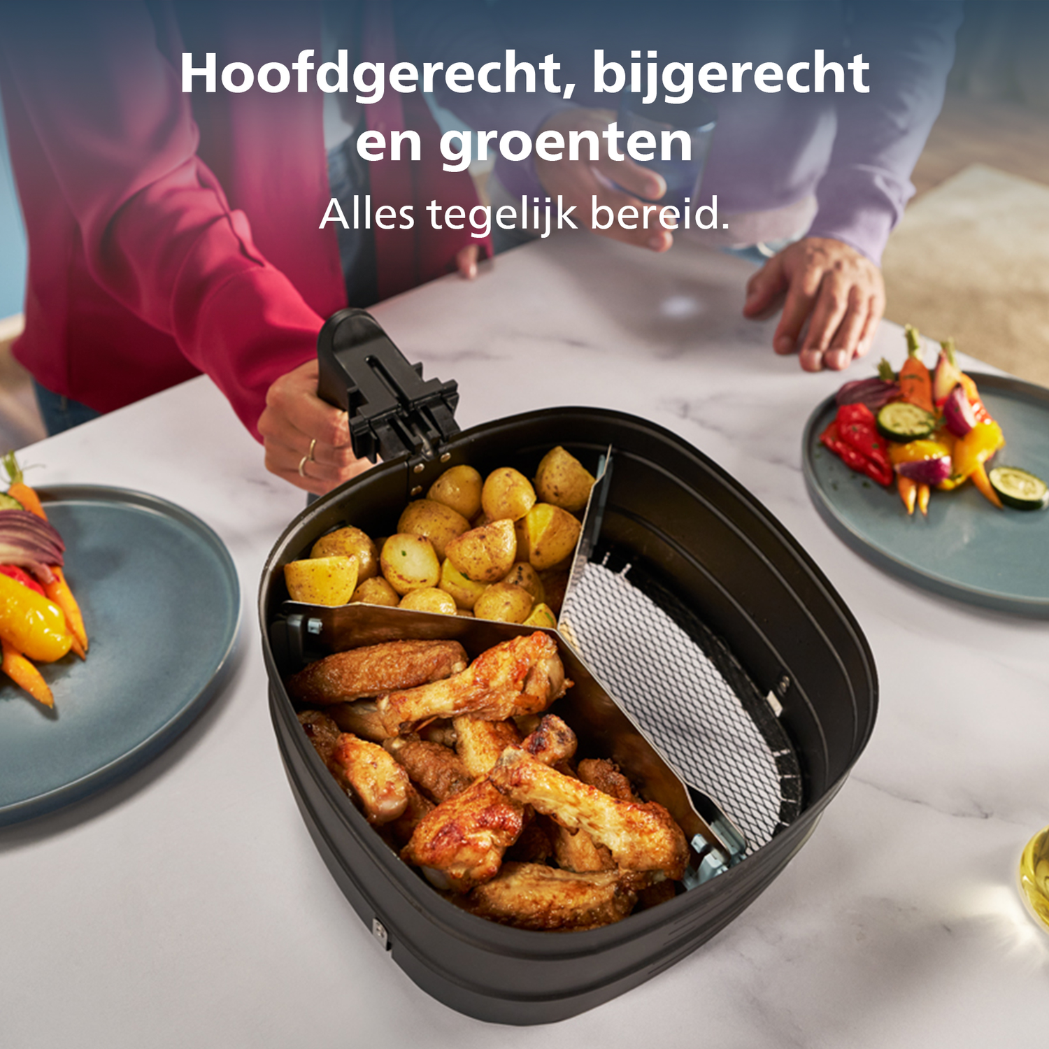 An air fryer with chicken wings, potatoes, and vegetables. Plates with grilled vegetables are on the sides. People are in the background.