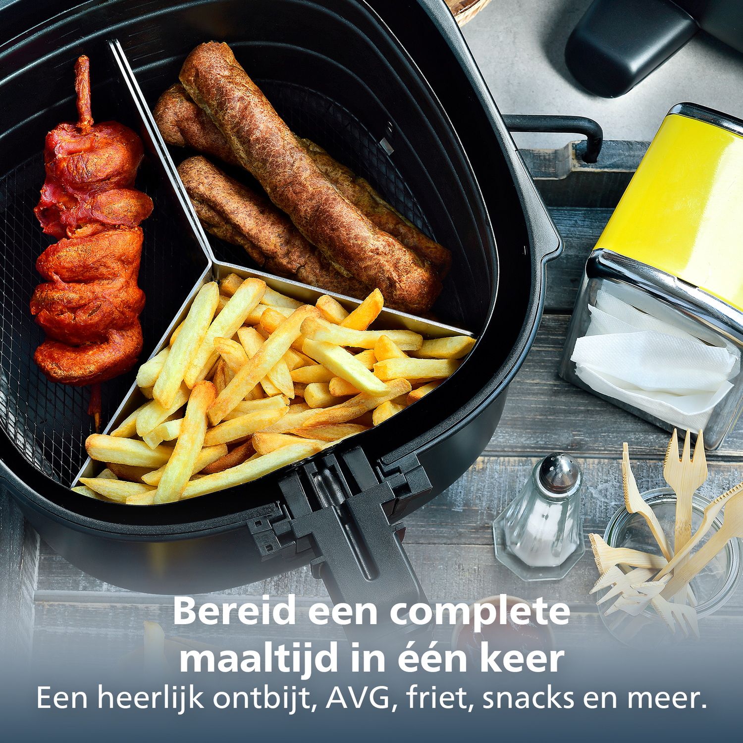 An air fryer with chicken skewers, sausages, and fries. A yellow napkin holder, salt shaker and wooden cutlery are on the side.