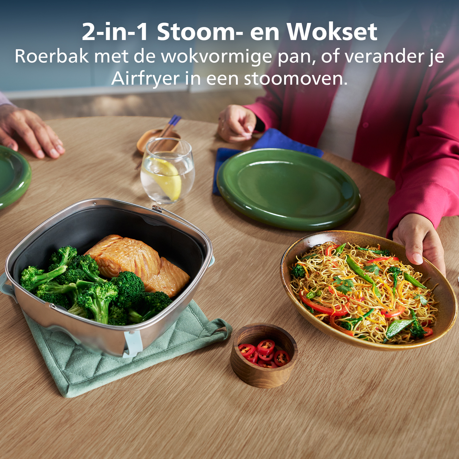 Een tafel met eten. Een stoomoven met zalm en broccoli. Een kom noodles wordt vastgehouden. Een kleine kom chilipepers.
