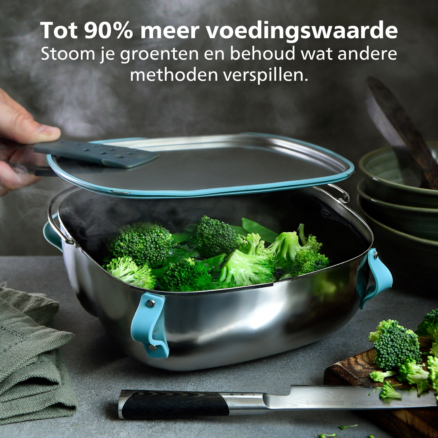 Een roestvrijstalen stoompan met broccoli, stoom stijgt op. Een persoon tilt het deksel op. Een mes en snijplank zijn in de buurt.