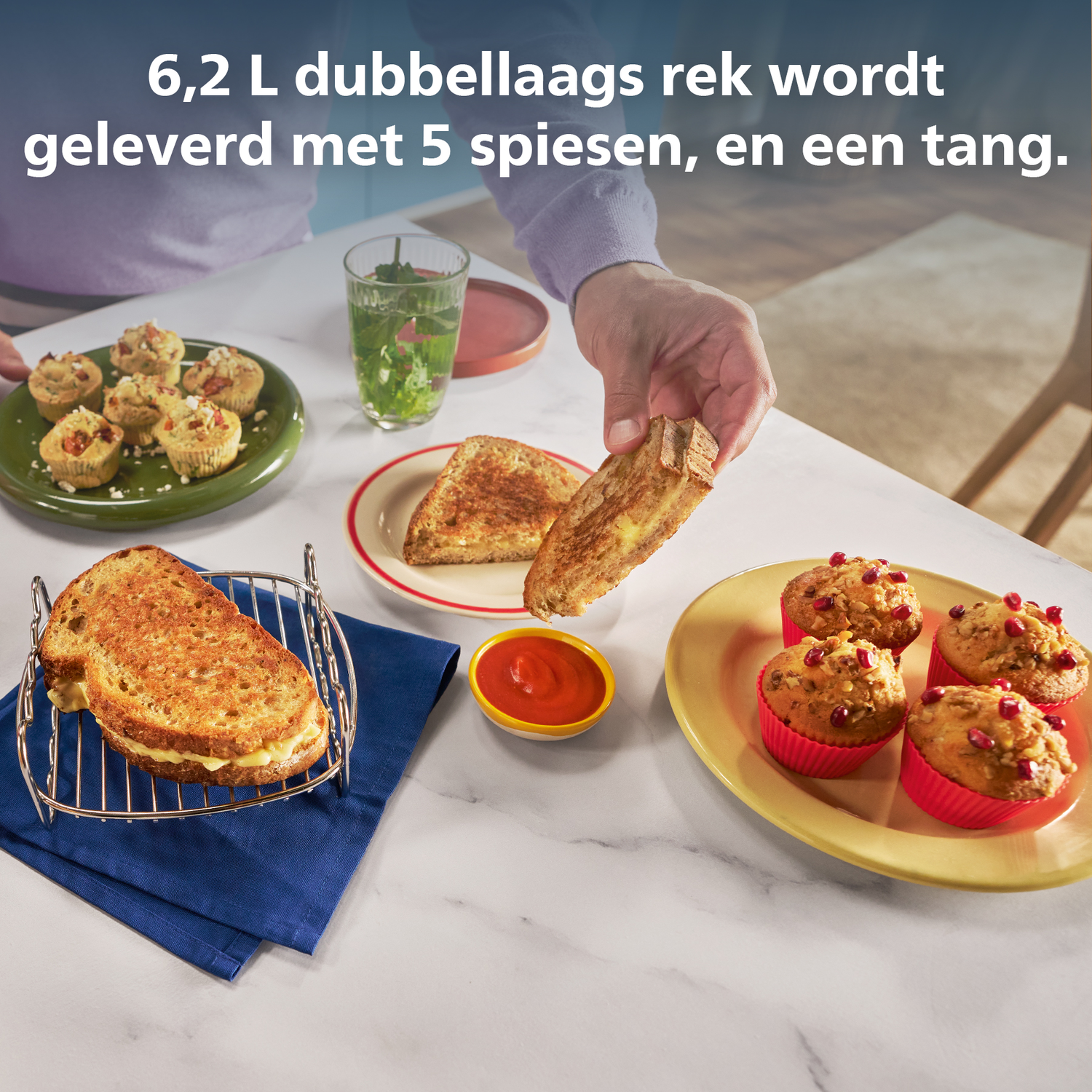 Een persoon houdt een gegrilde kaassandwich vast. Andere voedingsmiddelen zoals muffins en kleine gebakjes liggen op tafel.