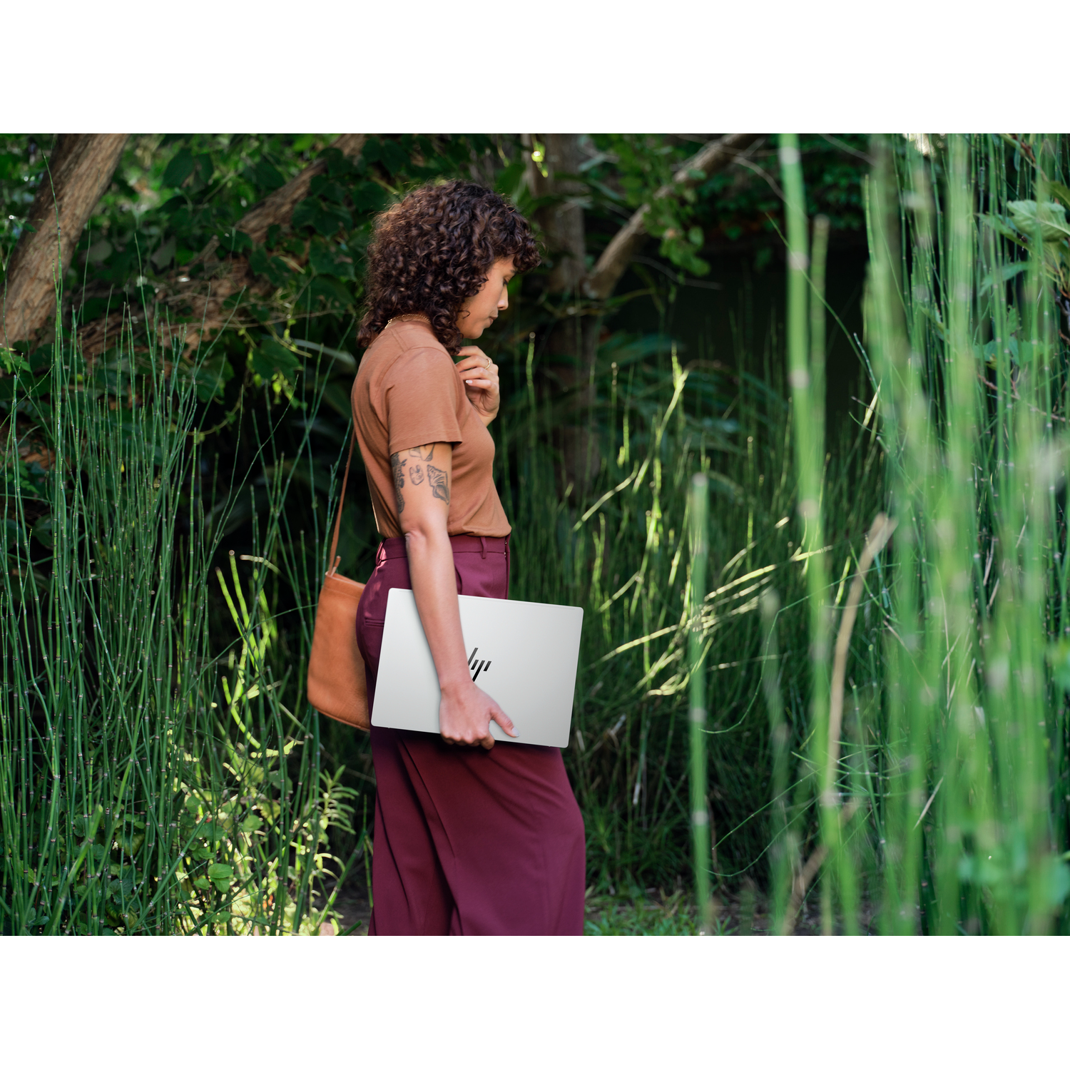 Femme en haut marron et pantalon bordeaux tient un ordinateur portable argenté, marchant dans la verdure avec un sac marron.