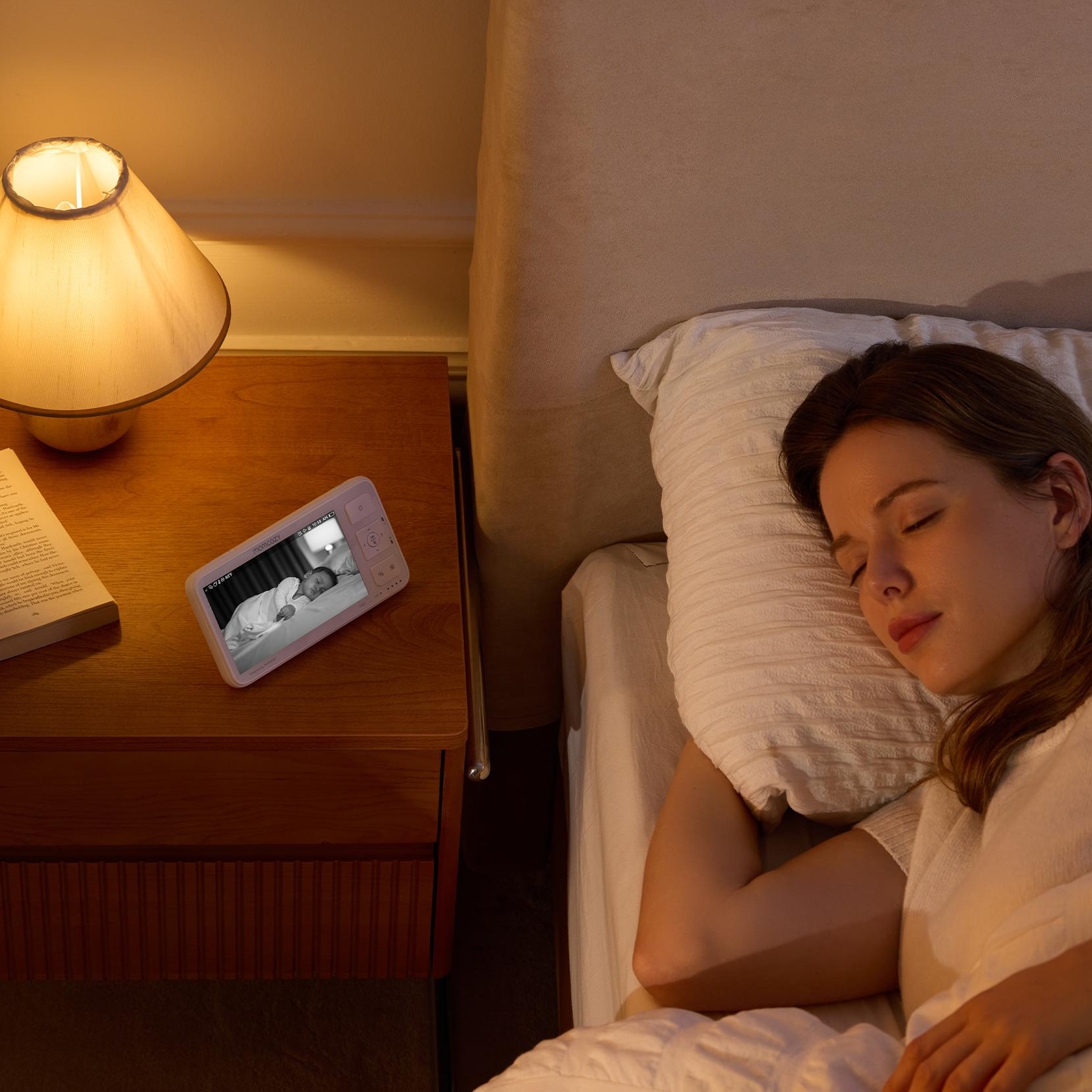 Eine Frau schläft im Bett. Ein Babyphone liegt auf einem Nachttisch, das ein Baby zeigt. Eine Lampe ist an.