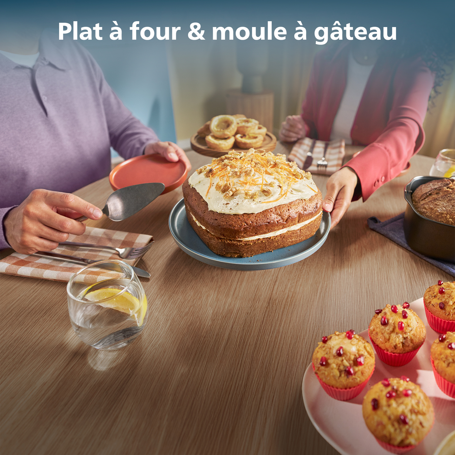 People at a table with a cake, muffins, and other baked goods. One person holds a cake server, another a plate. 