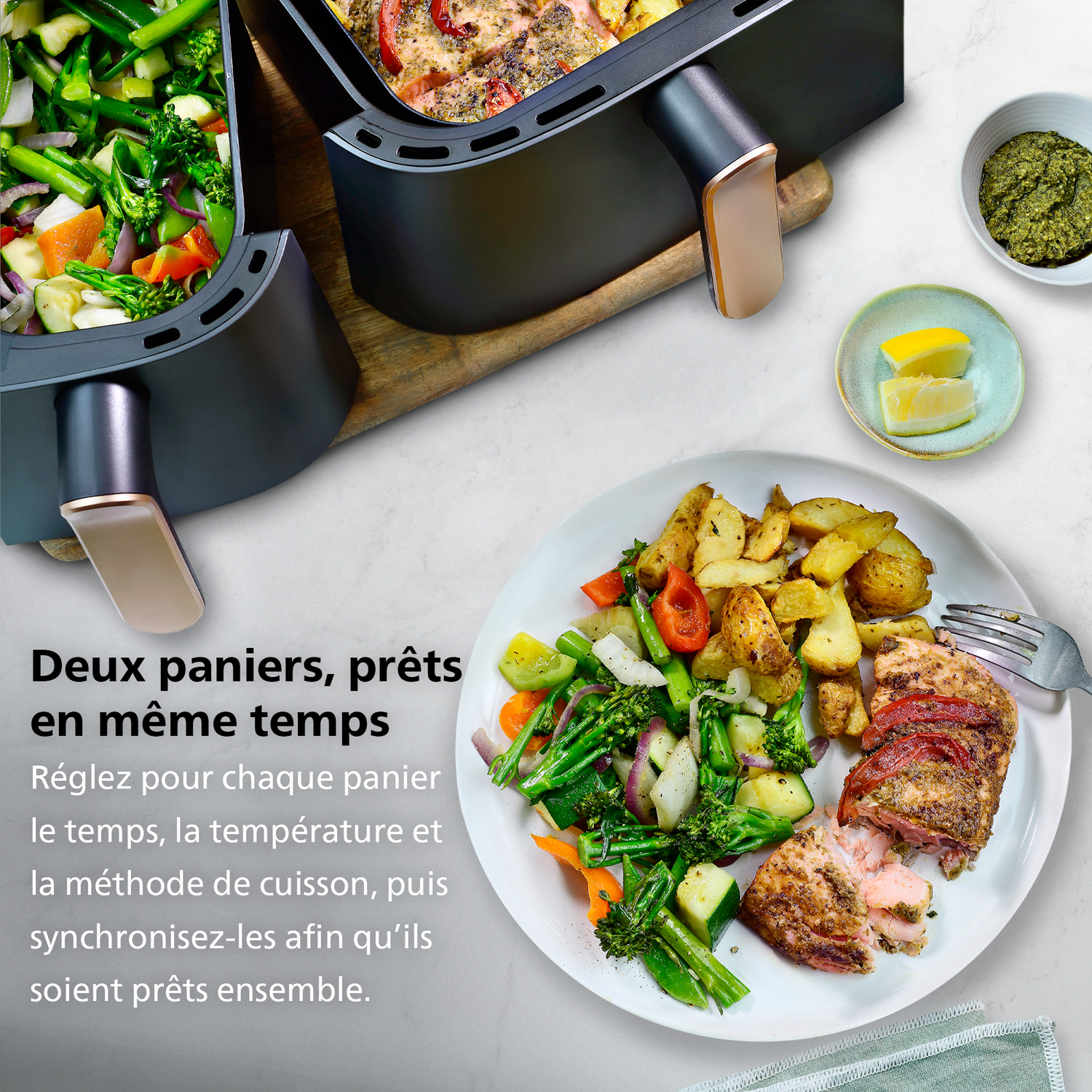 Une friteuse à air avec deux paniers, l'un avec des légumes, l'autre avec de la nourriture. Une assiette contient de la nourriture, des tranches de citron et du pesto.