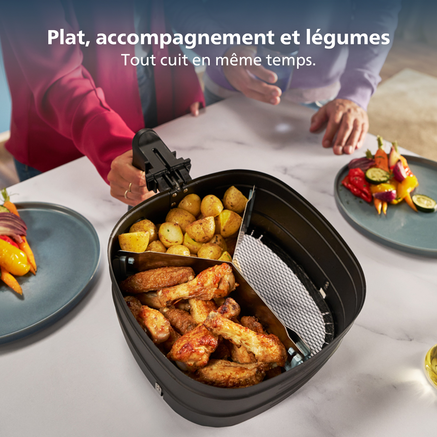 An air fryer with potatoes, chicken wings, and vegetables. Two people stand over the fryer. Plates with food are on each side.