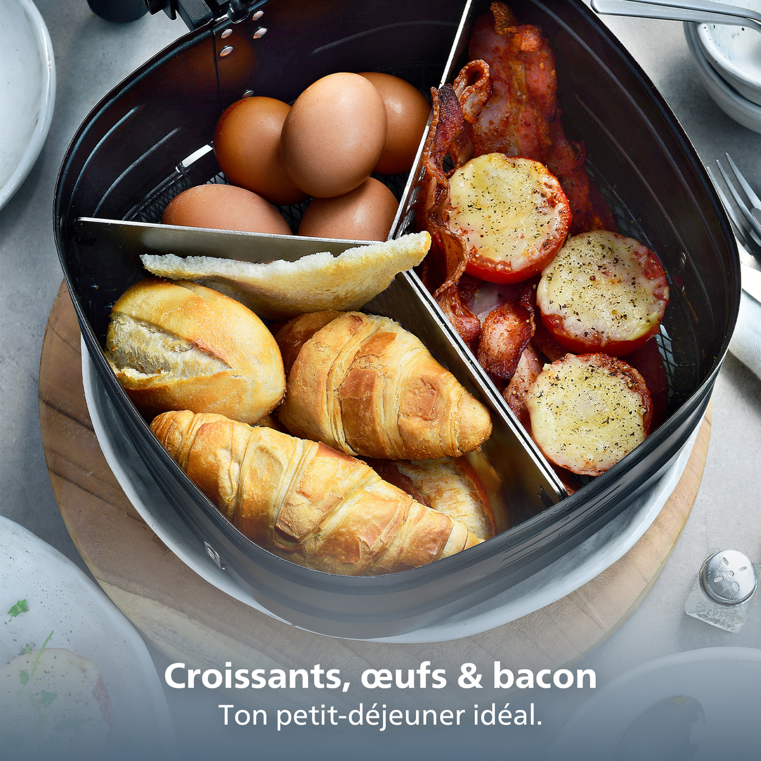 Breakfast contains eggs, bacon, tomatoes, croissants, and bread rolls in a black container. The food is on a wooden board.