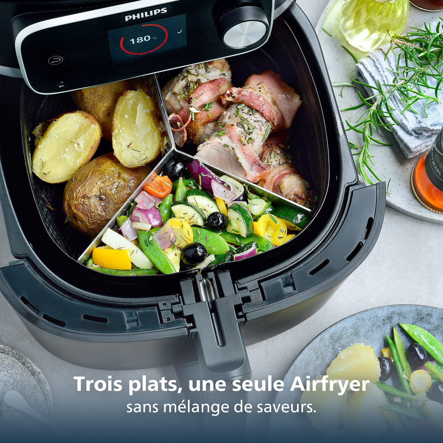 An air fryer with potatoes, bacon-wrapped meat, and vegetables. The display shows 180°C. Text says 'Three dishes, one Airfryer'.