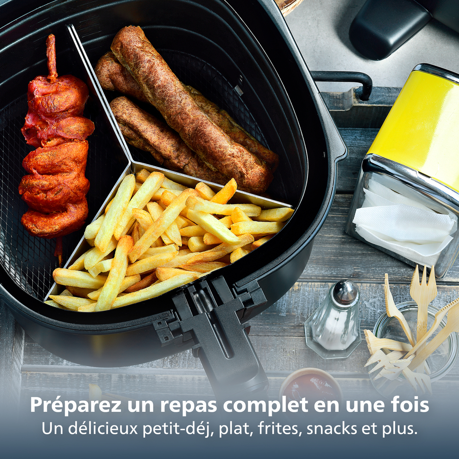 An air fryer with food inside: chicken skewers, sausages, and fries. A yellow napkin holder is on the right, with wooden forks.