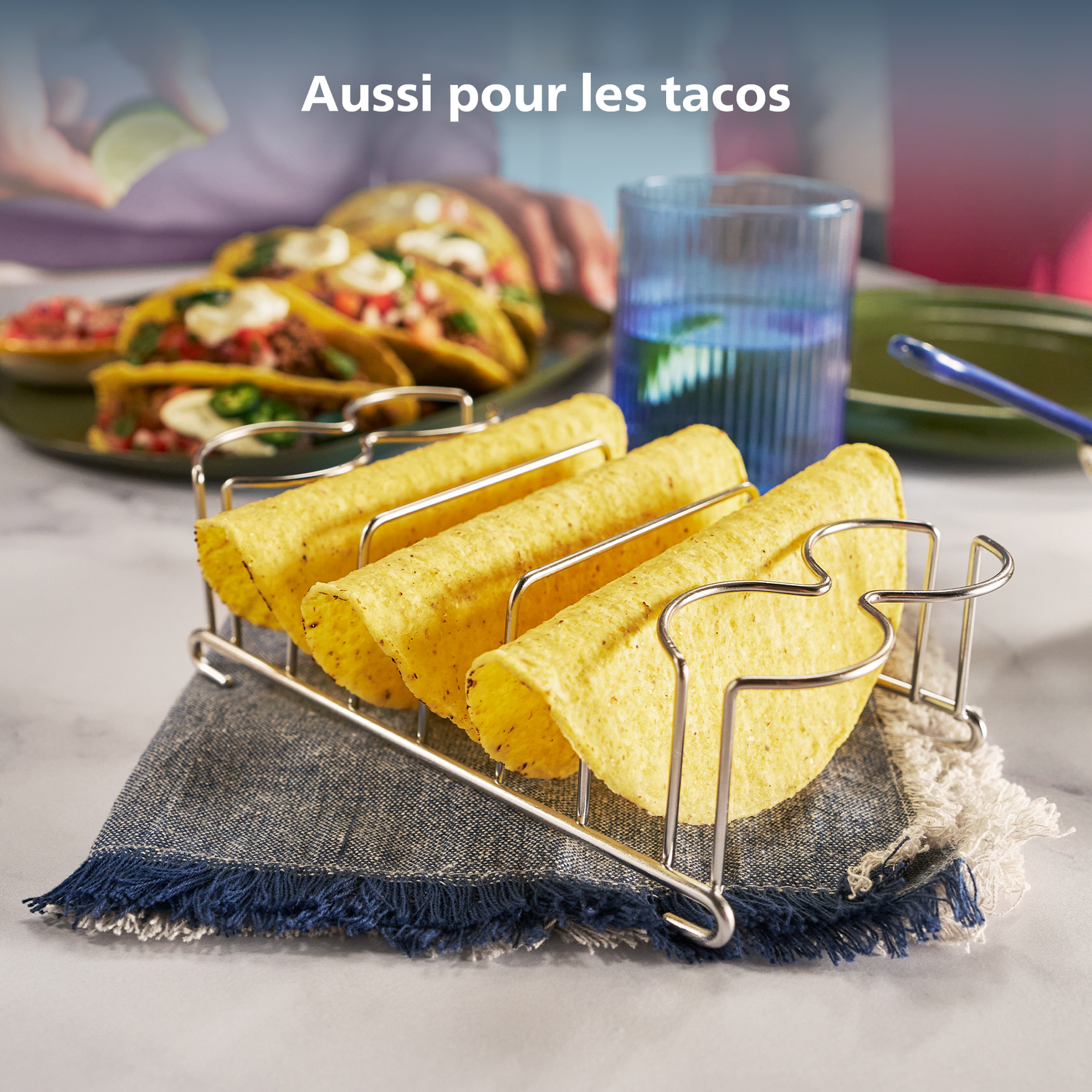 Gele tacoschelpen in een metalen houder op een blauwe servet. Tacos met vulling op de achtergrond, met een blauw glas en limoen.