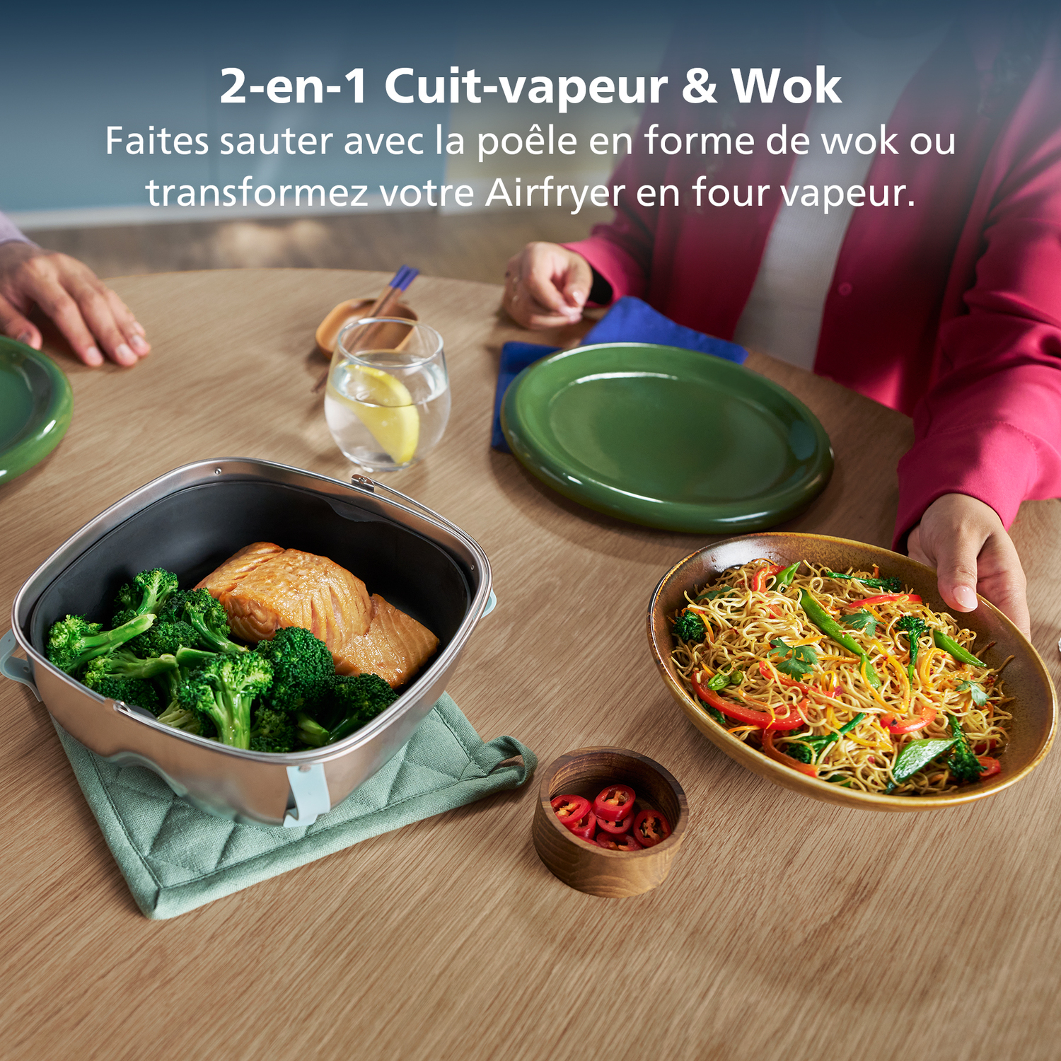 Een tafel met eten: broccoli en zalm, noedels en chilipepers. Een persoon houdt een bord noedels vast.