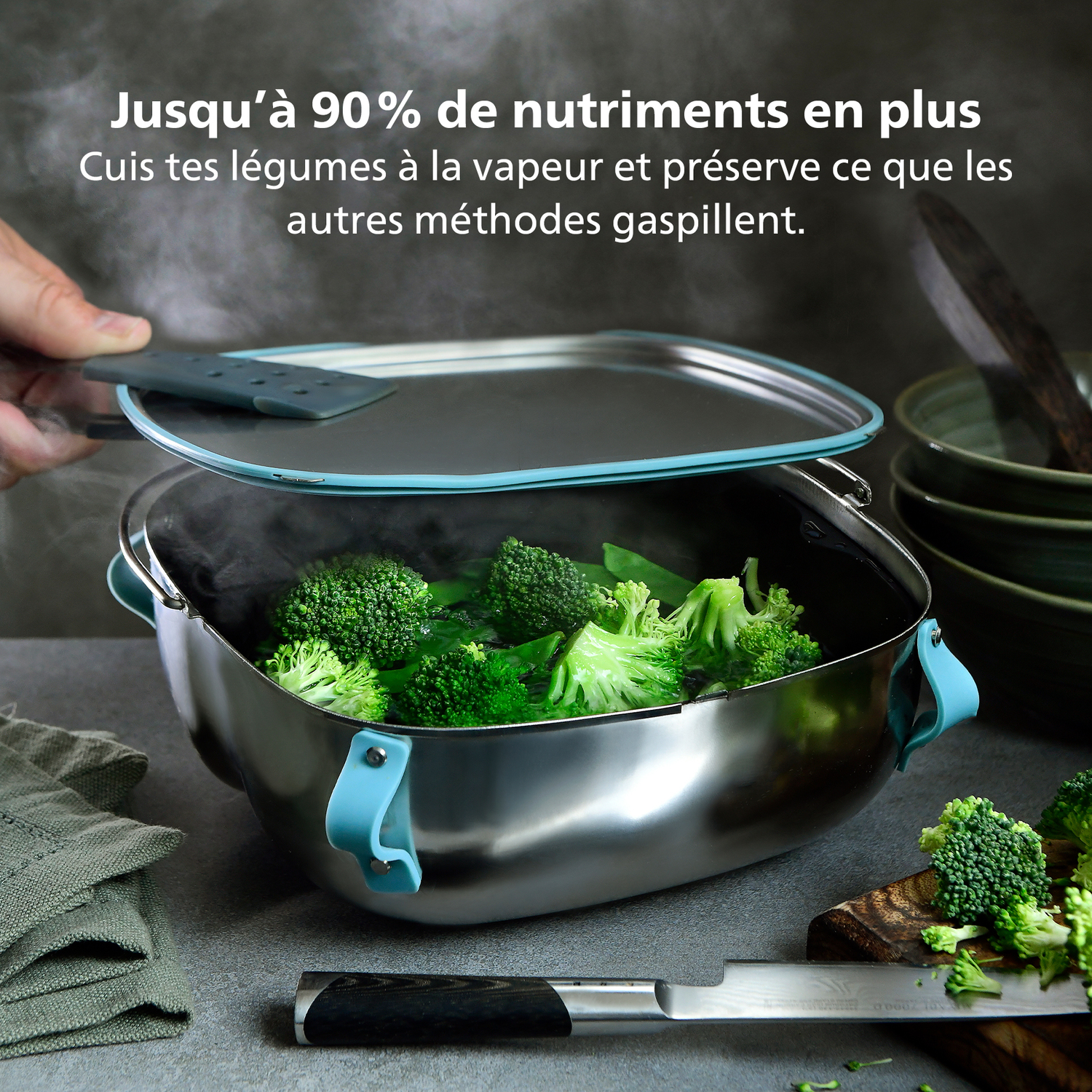 Een persoon tilt het deksel op van een stomende pan met broccoli. De pan is van roestvrij staal met blauwe accenten. Mes en houten snijplank zijn aanwezig.